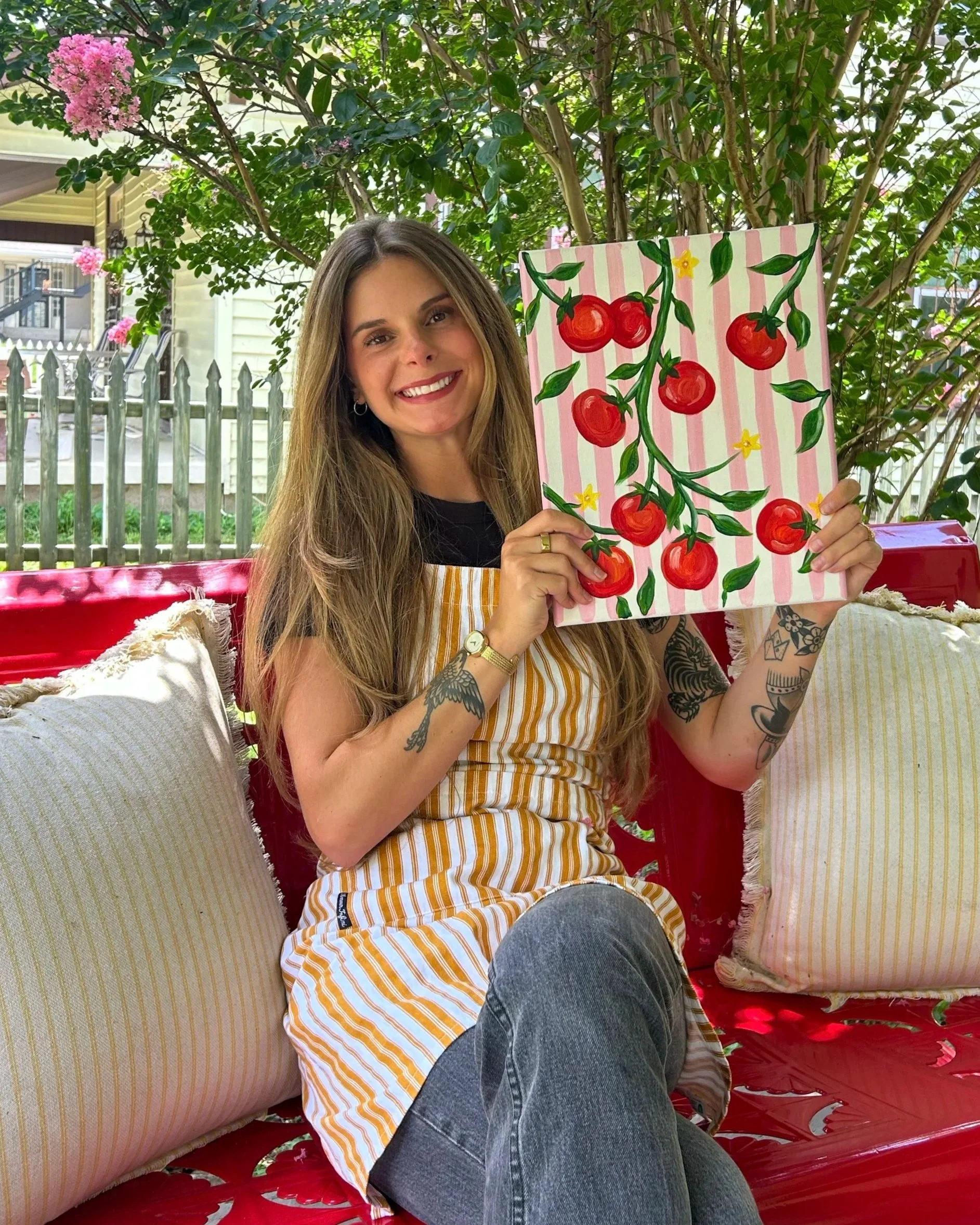 Girl with blonde hair on a red bench outside holding a painting with stripes and tomatoes