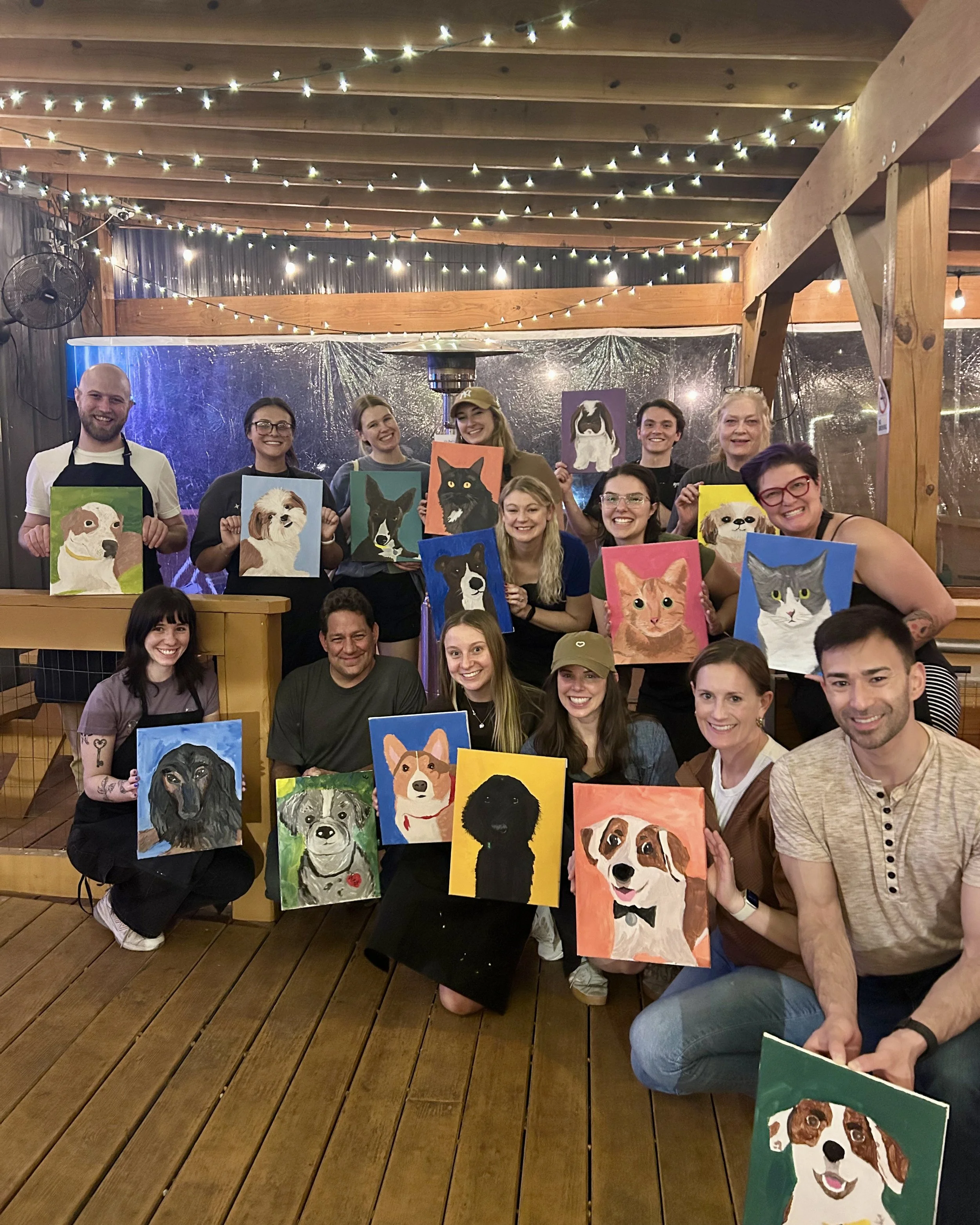 Group of people holding colorful paintings of their pets under string lights