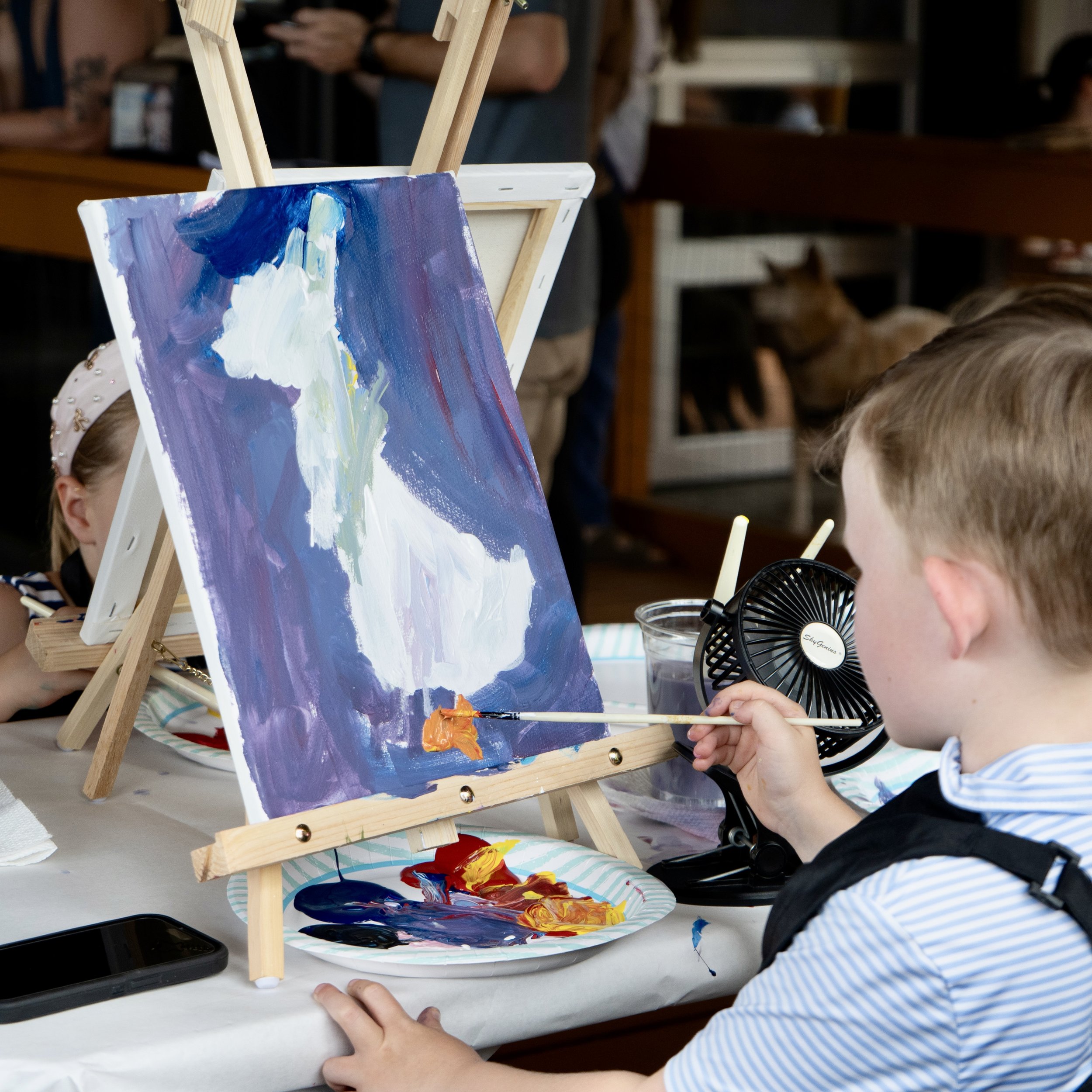 A young boy with blonde hair paints a purple canvas with a white bird outside