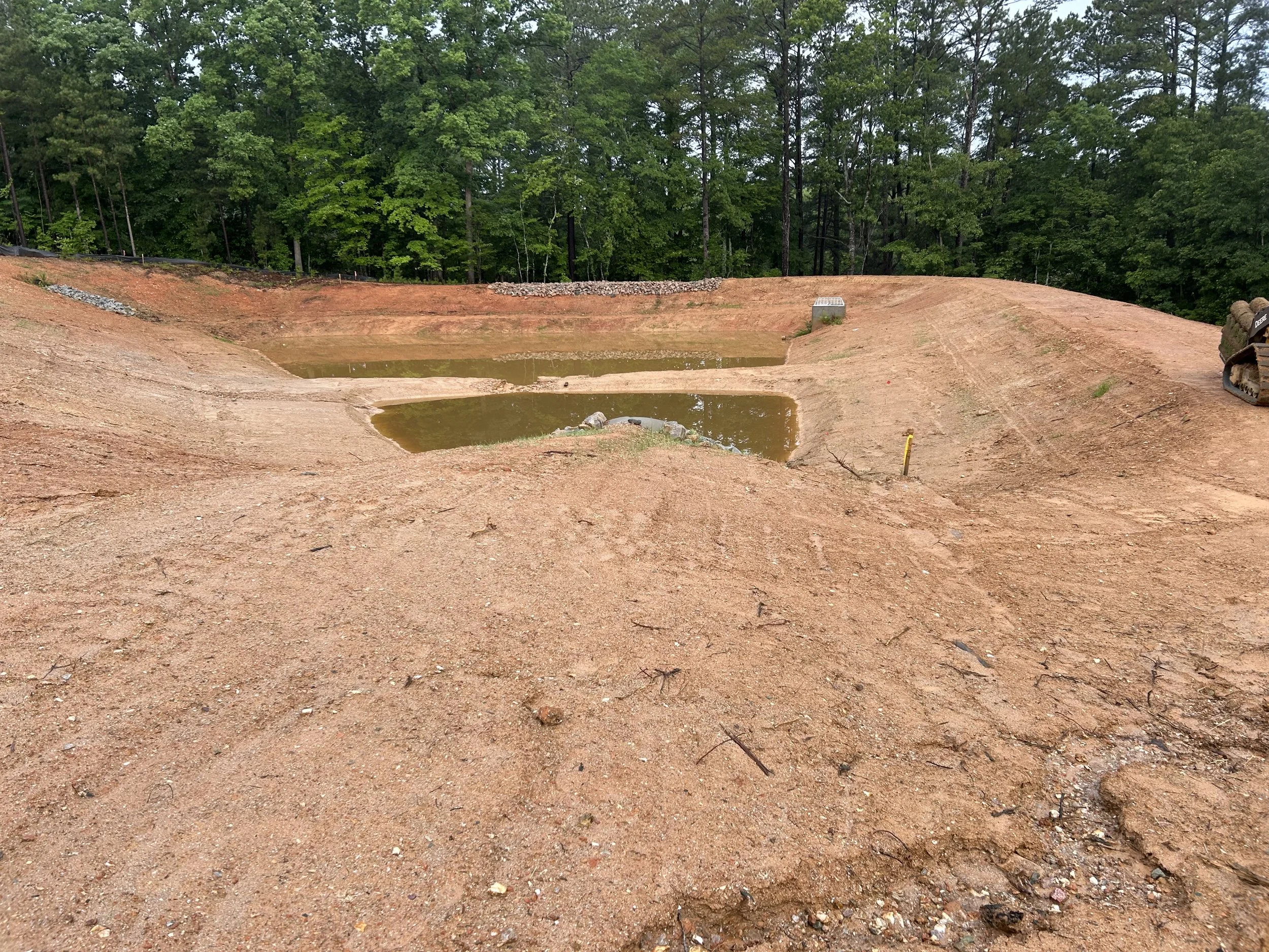 Construction site with excavated area and small pools of water, surrounded by dirt and green trees in the background.