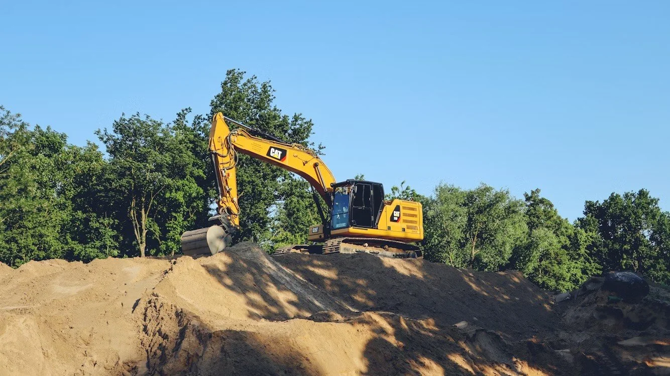 A yellow CAT excavator working on a sandy construction site with trees in the background.