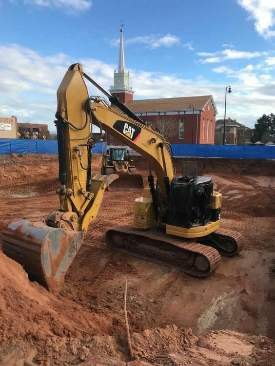 A small CAT excavator working on a construction site with a church and blue sky in the background.