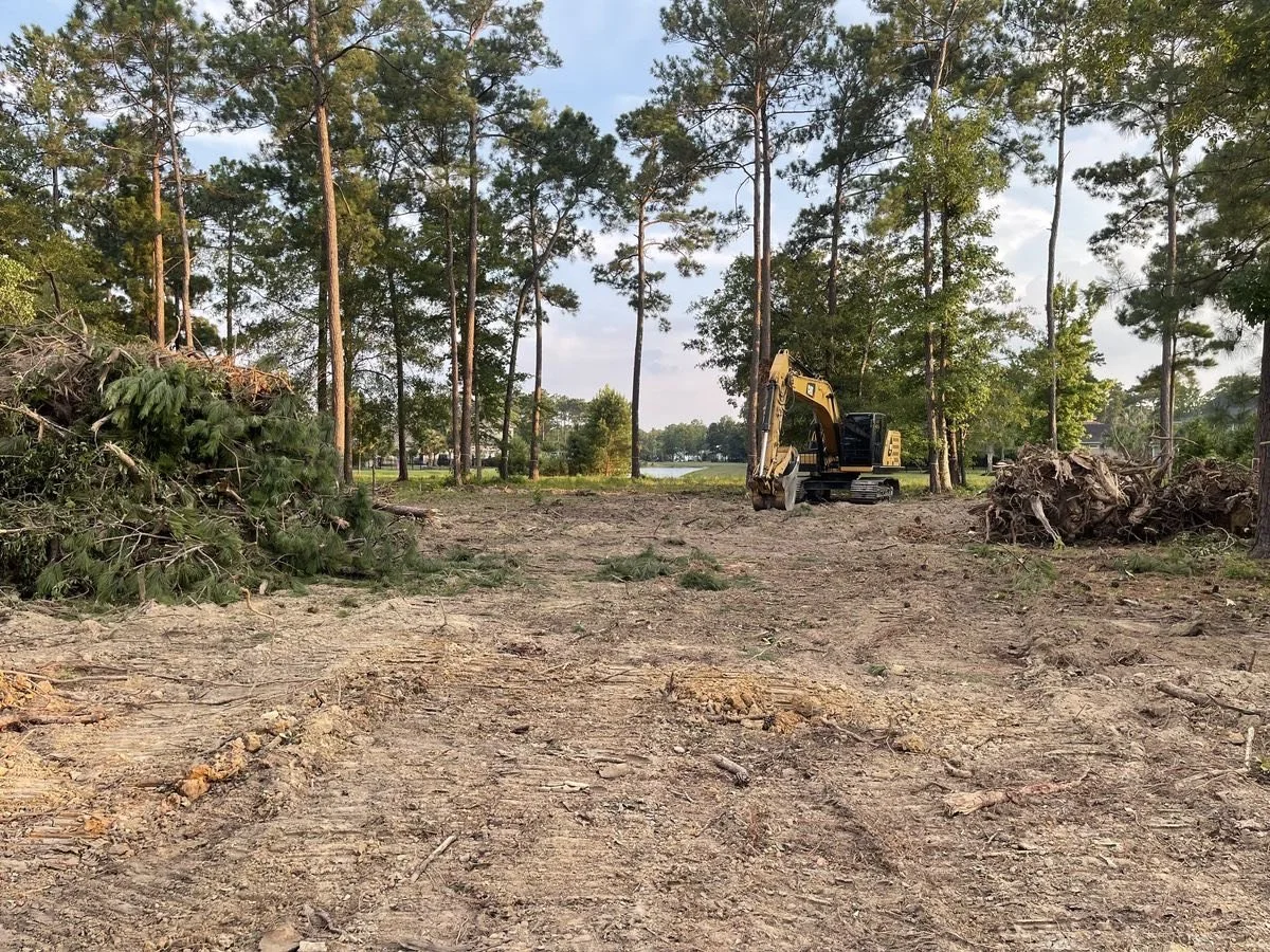A small excavator working on a cleared area of land with several fallen trees and forest in the background.