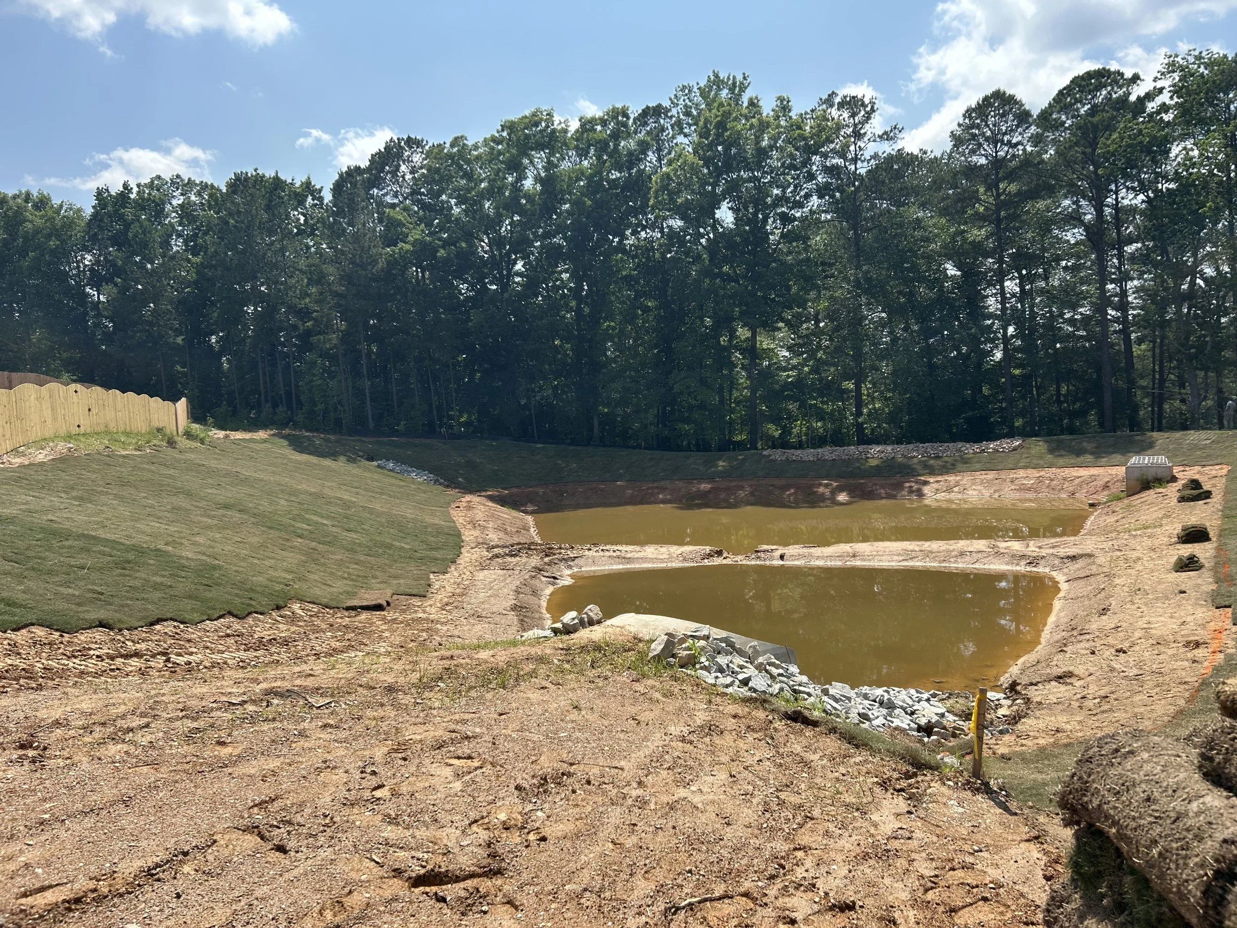 Construction site with two dirt-filled ponds, Sod install on one side, and a wooded area in the background.
