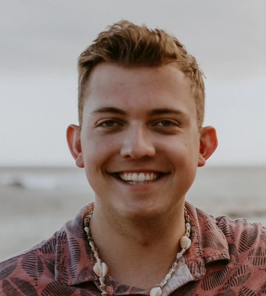 A smiling young man with short, light brown hair, wearing a patterned shirt and shell necklace, standing on a beach with the ocean in the background.