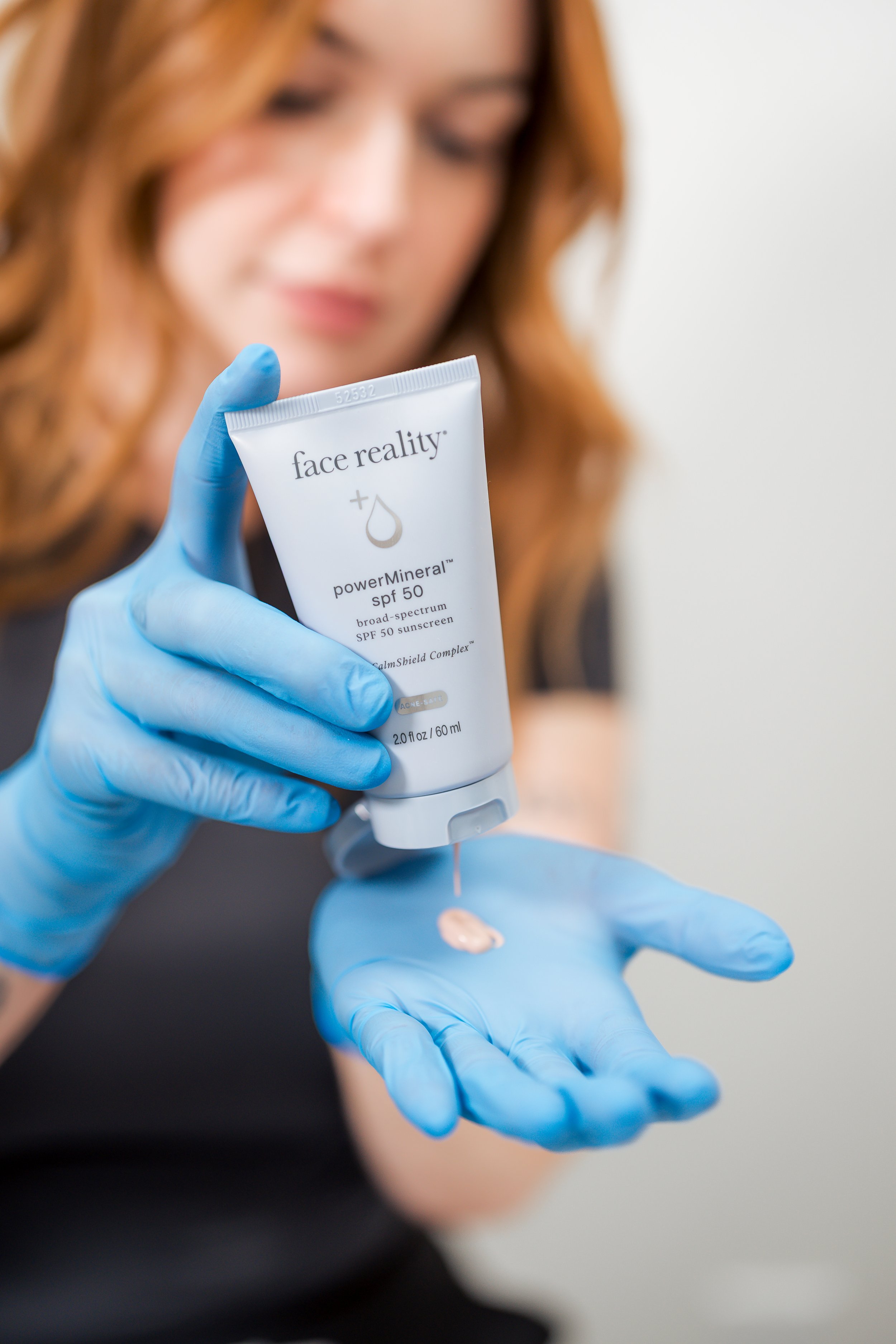 A woman with light skin and curly red hair is applying lotion from a tube labeled 'face reality' with SPF 50. She is wearing blue disposable gloves and appears to be in a skincare or medical setting.