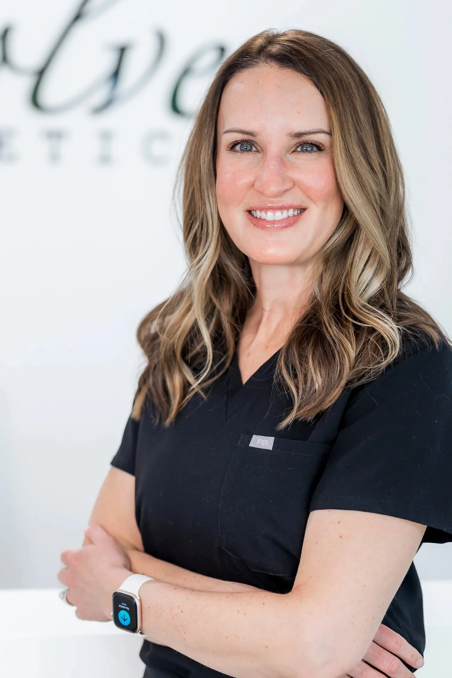 A woman with shoulder-length wavy light brown hair wearing a black top with a nametag reading 'FIGS' and a small gold necklace, smiling against a plain white background.
