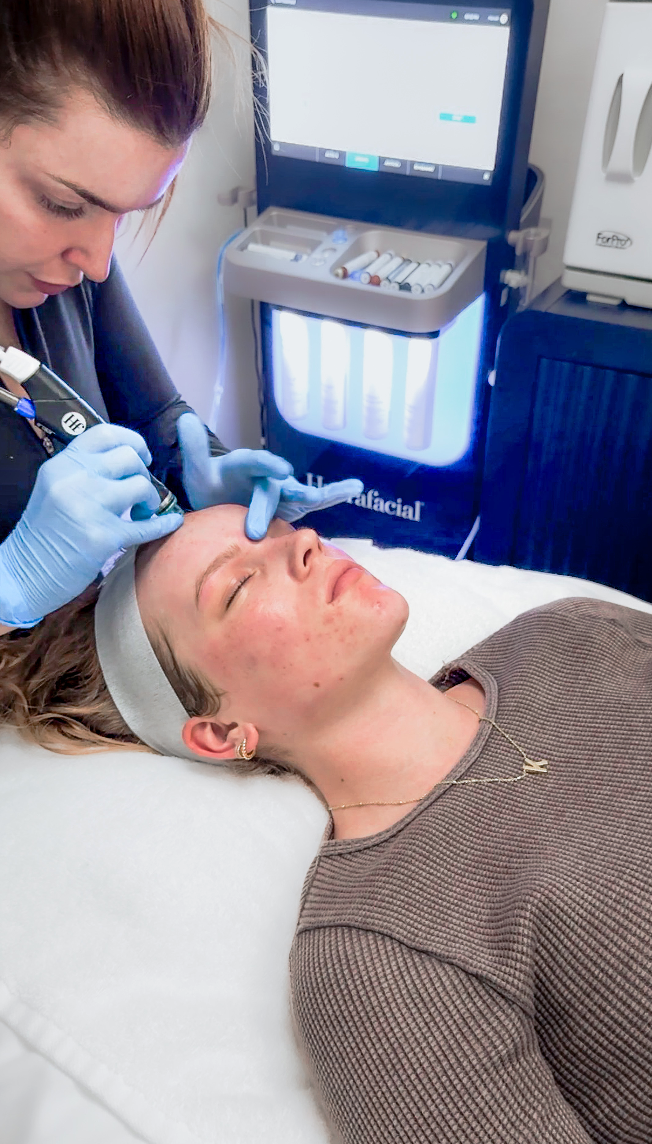 A woman is lying on a medical bed with her eyes closed while a healthcare professional, wearing gloves, performs a cosmetic or dermatological procedure on her forehead. In the background, there's medical equipment and a computer screen.