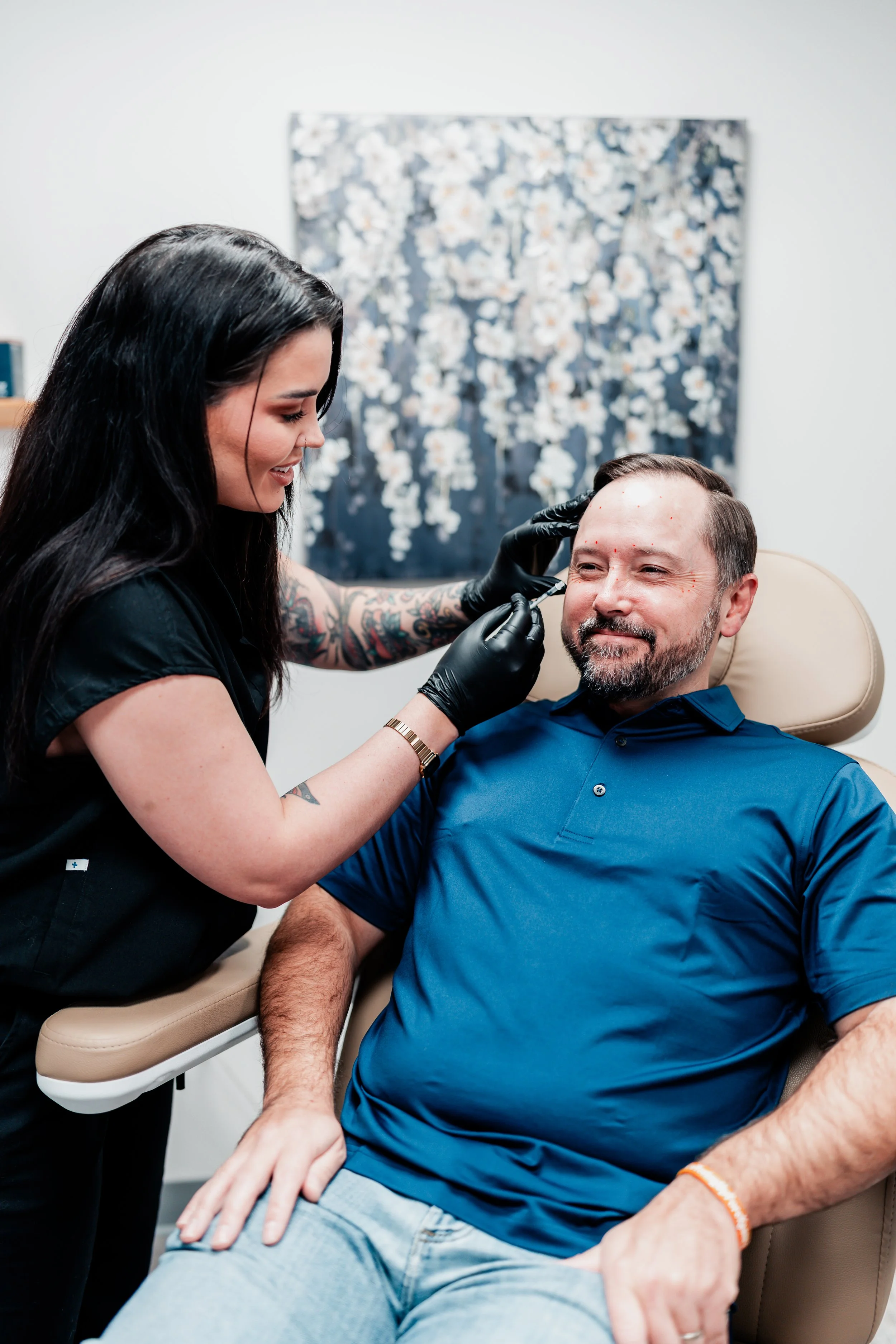 A female healthcare professional with tattoos on her right arm is administering a cosmetic or medical injection to a middle-aged man with a beard, sitting in a medical chair. The man is smiling and wearing a blue polo shirt.