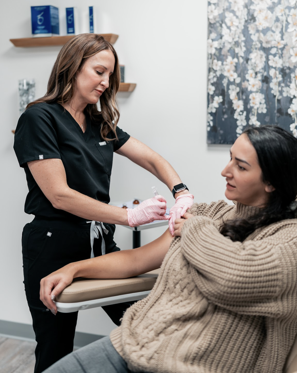 A nurse is administering an injection to a woman reclined on a medical chair in a clinic or medical office, with shelves and artwork in the background.