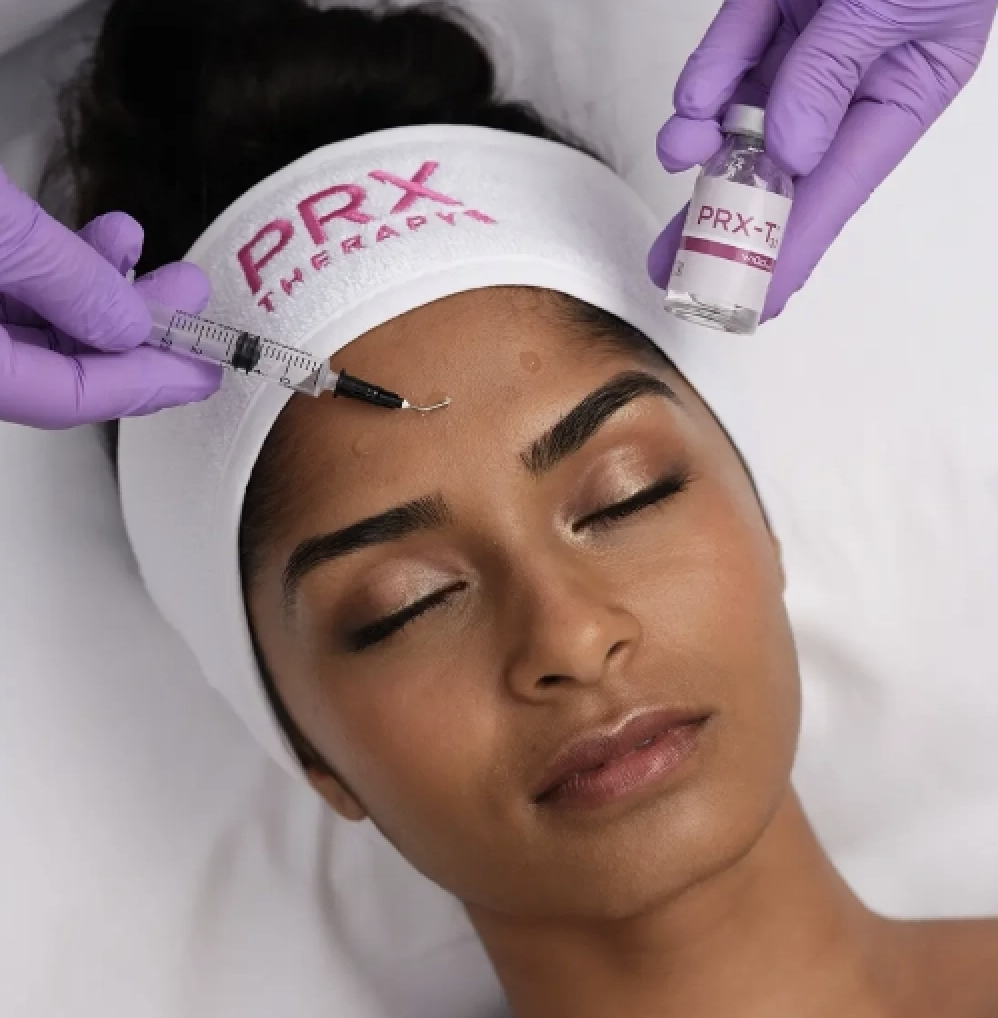 A woman receiving a skincare treatment on her forehead with a syringe from a professional wearing purple gloves, while lying down with a white headband that says 'PRX THE TREAT!'