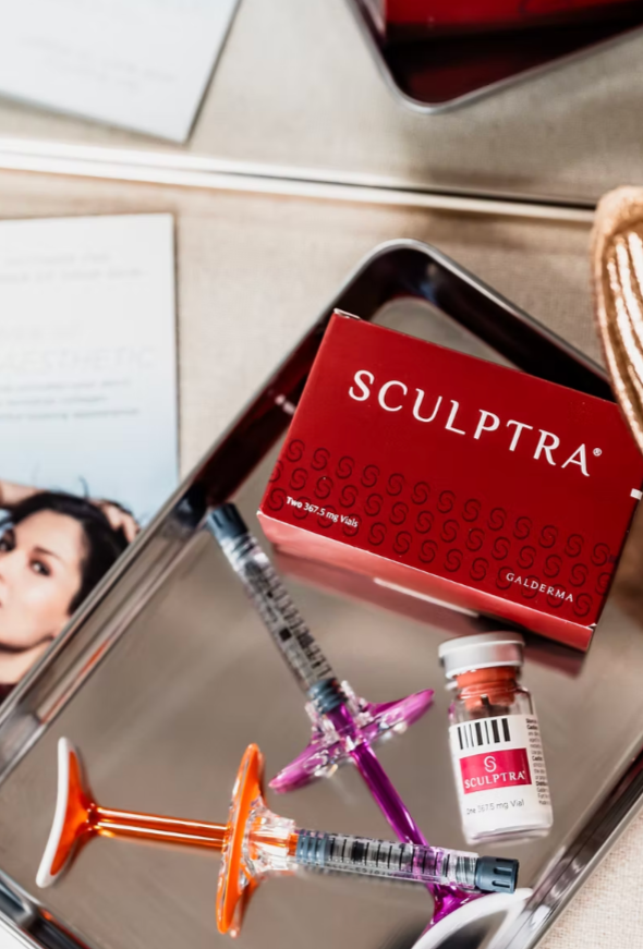 Sculptra cosmetic treatment products including a red box labeled 'Sculptra', two syringes with orange and purple caps, and a small vial with a barcode and label, placed on a reflective metallic tray.