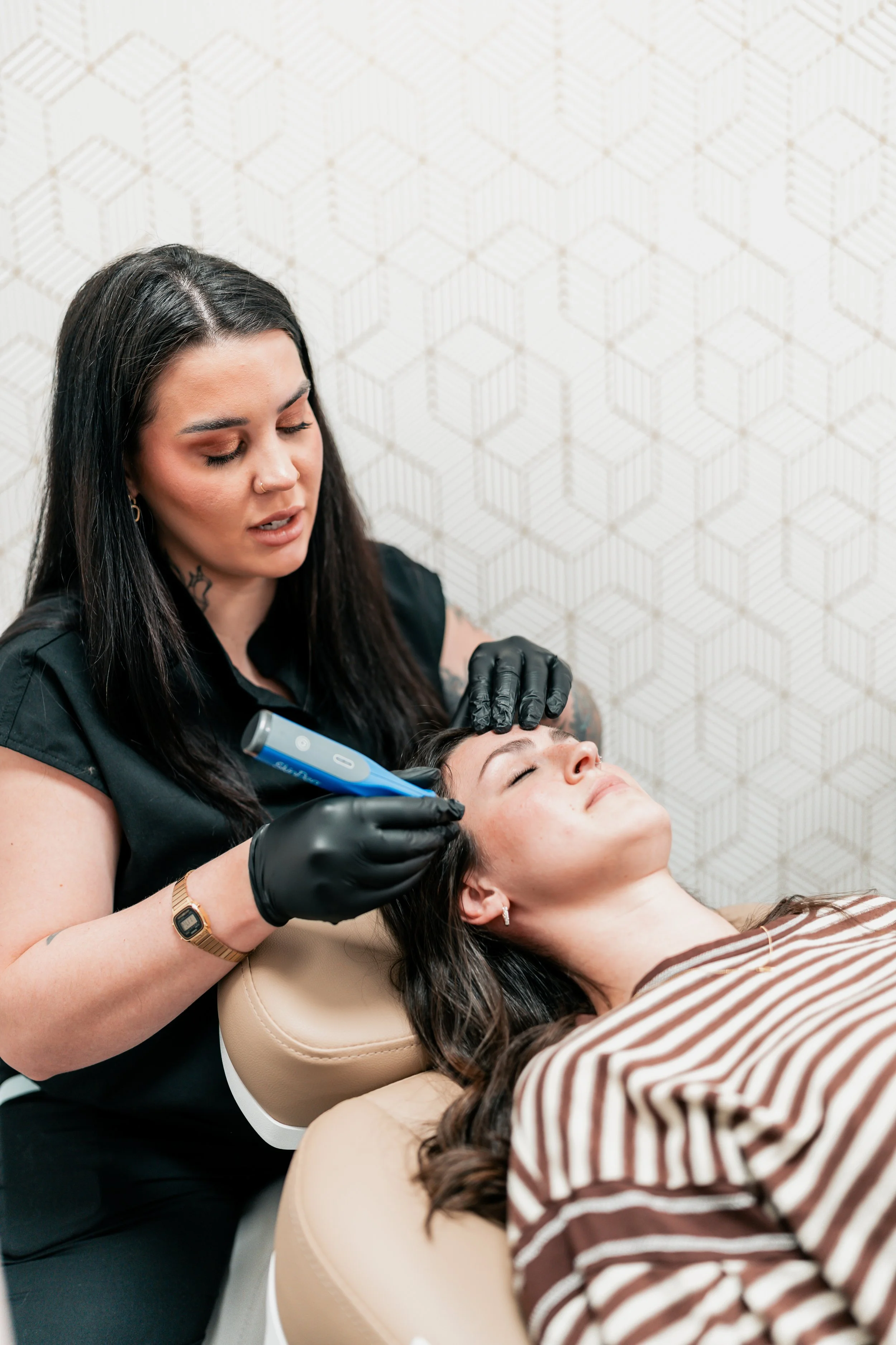 A woman lying on a treatment chair receiving a facial filler treatment from a cosmetologist using a handheld device.