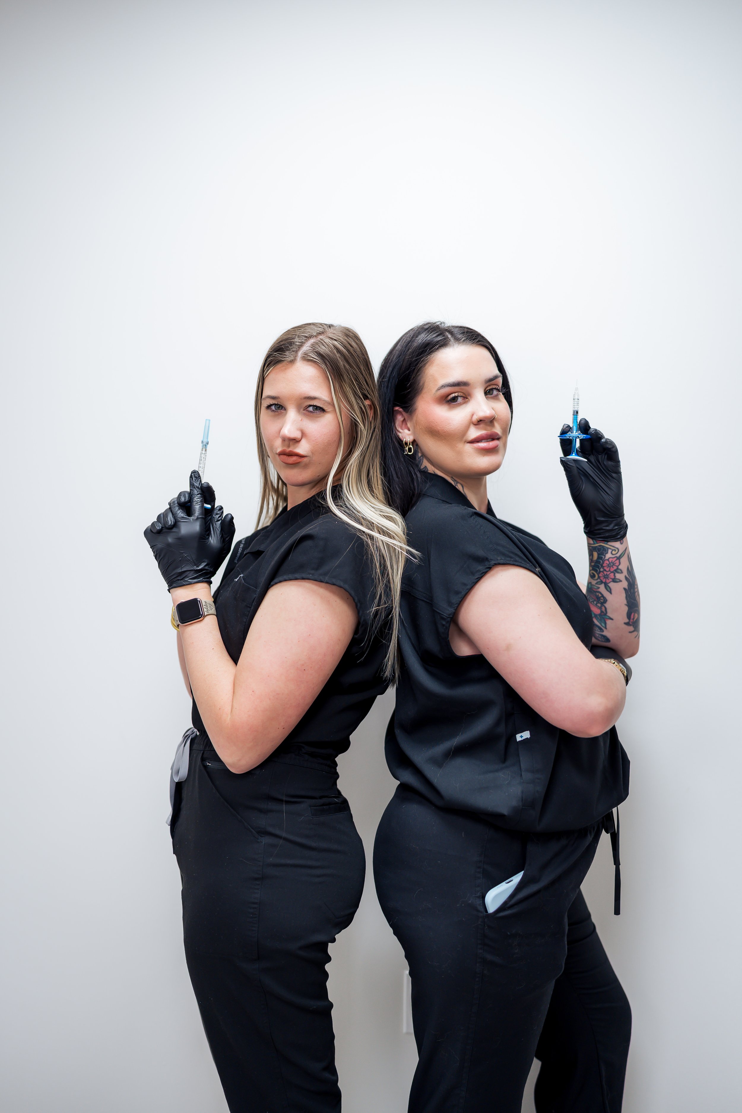 Two women posing back-to-back, each holding a syringe with botox, daxxify, and dysport neurotoxins, wearing black medical scrubs and gloves in front of a plain white background.