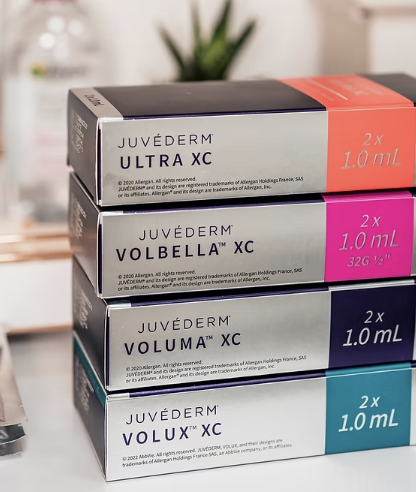 Stack of four boxes of Juvéderm dermal fillers in different colors for different formulations, placed on a reflective surface with a blurred plant and water bottle in background.