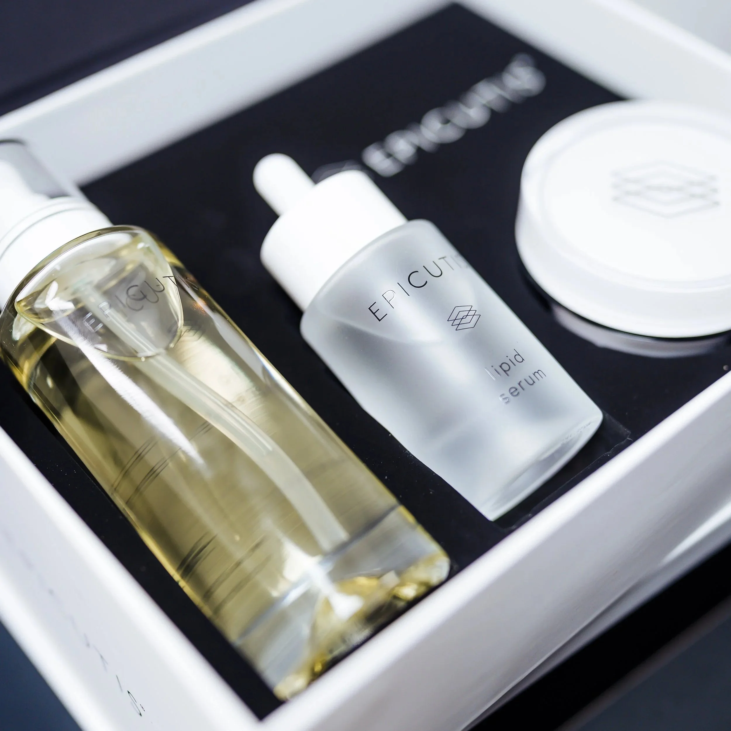 A skincare gift set with a clear serum bottle, a white dropper bottle labeled 'Epicuutis Lipid Serum,' and a white jar with a lid, all arranged in a black box.
