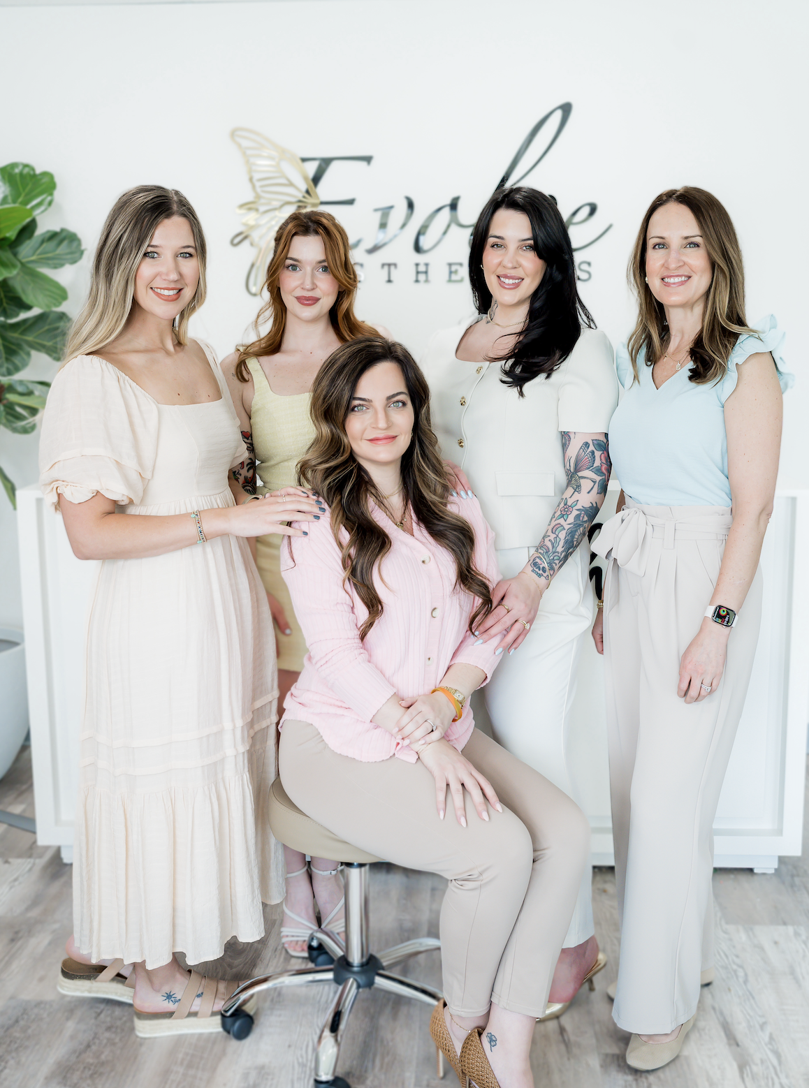 Group of five women posing together in front of a sign that reads 'Evolve,' with an abstract butterfly design, in a bright, modern interior.