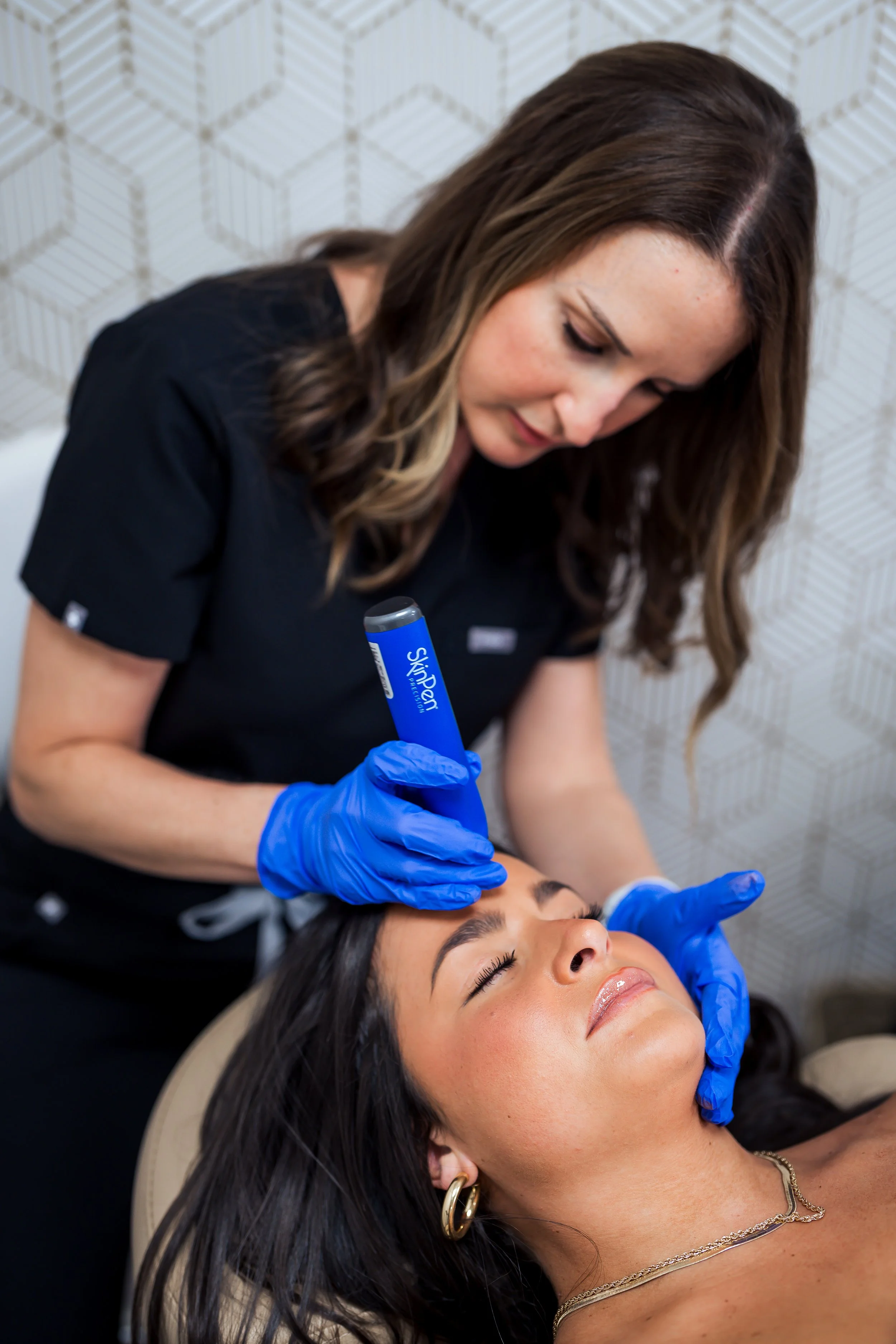 A woman lying down with her eyes closed and a slight smile receives a cosmetic treatment to her forehead by a professional wearing blue gloves, with another person partially visible in the background.