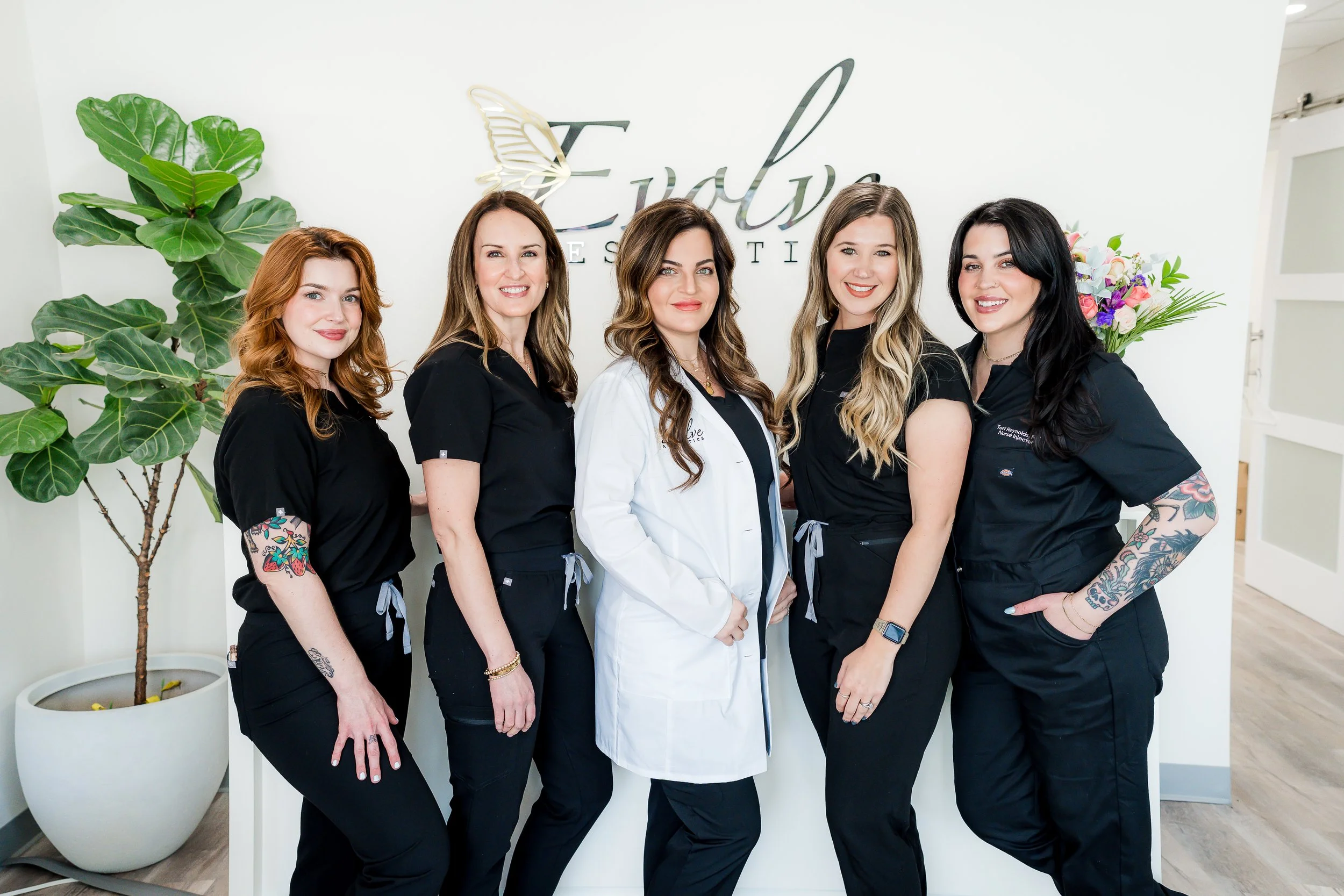 Five women standing side by side indoors, smiling, with a white wall and plants in the background.