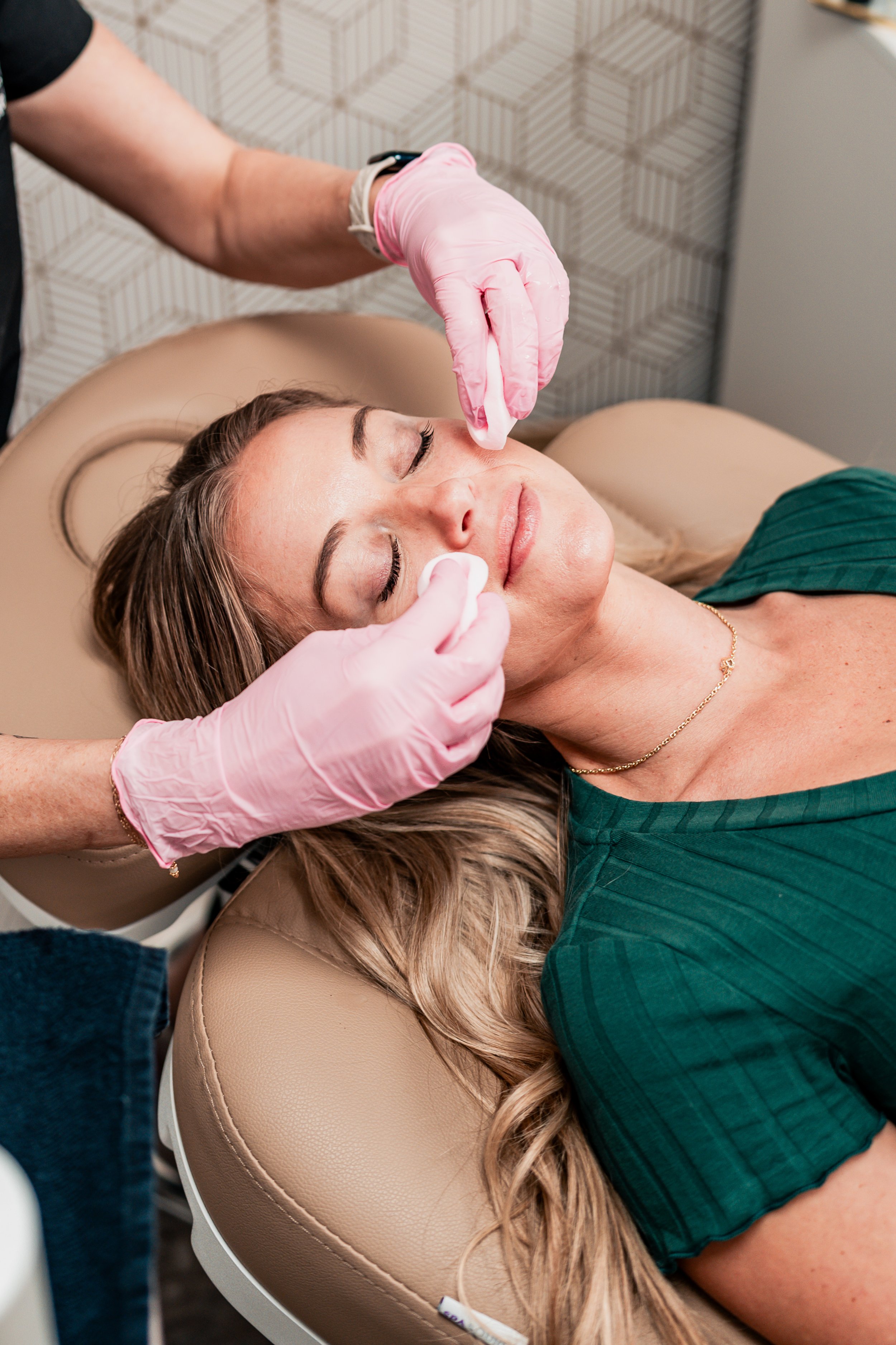 A woman lying on a treatment bed with eyes closed, receiving a facial treatment from a skincare professional wearing pink gloves.