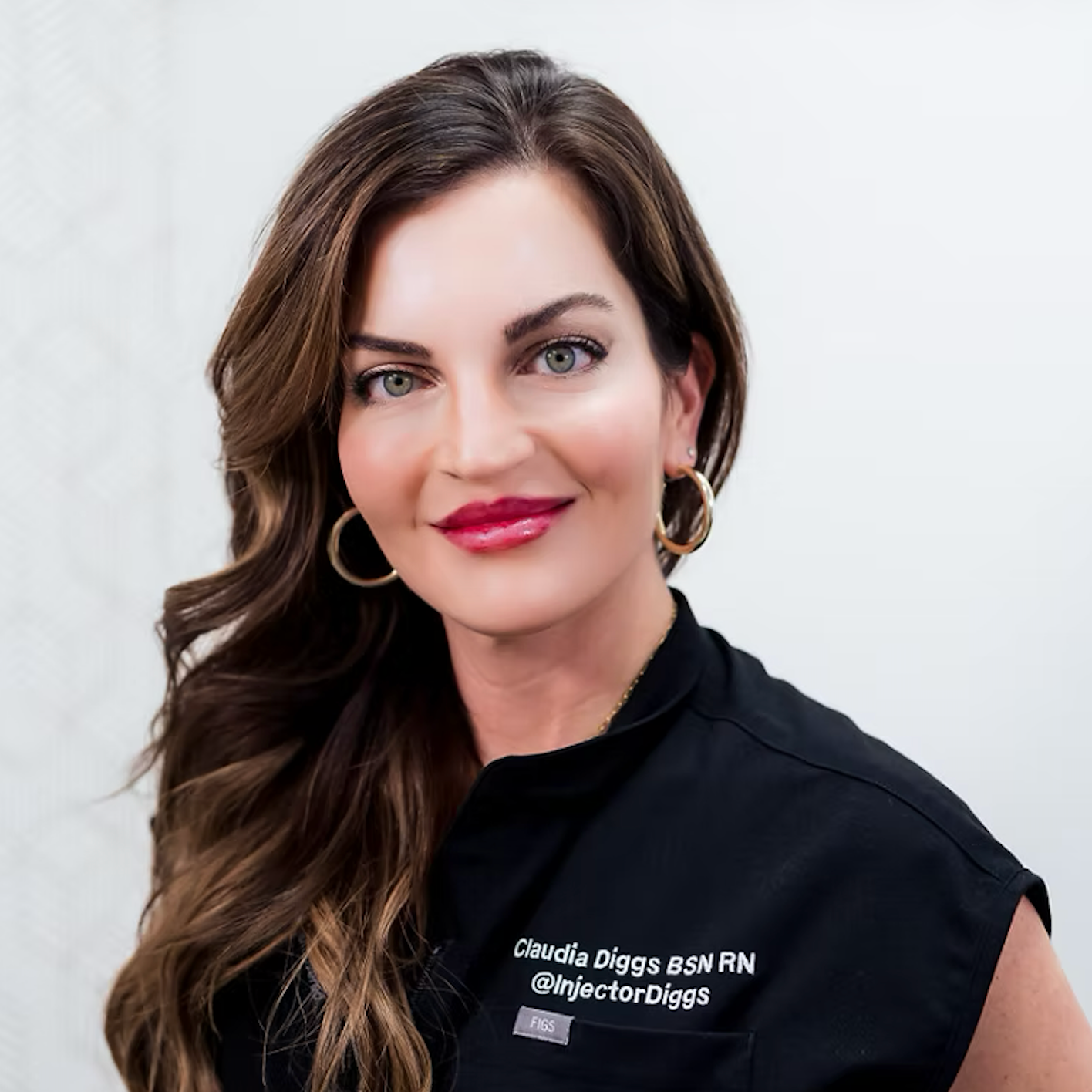 Portrait of a woman with long wavy brown hair, wearing hoop earrings, and a black shirt with embroidered text reading 'Claudia Diggs BSN RN @injectorDiggs,' against a plain white background.