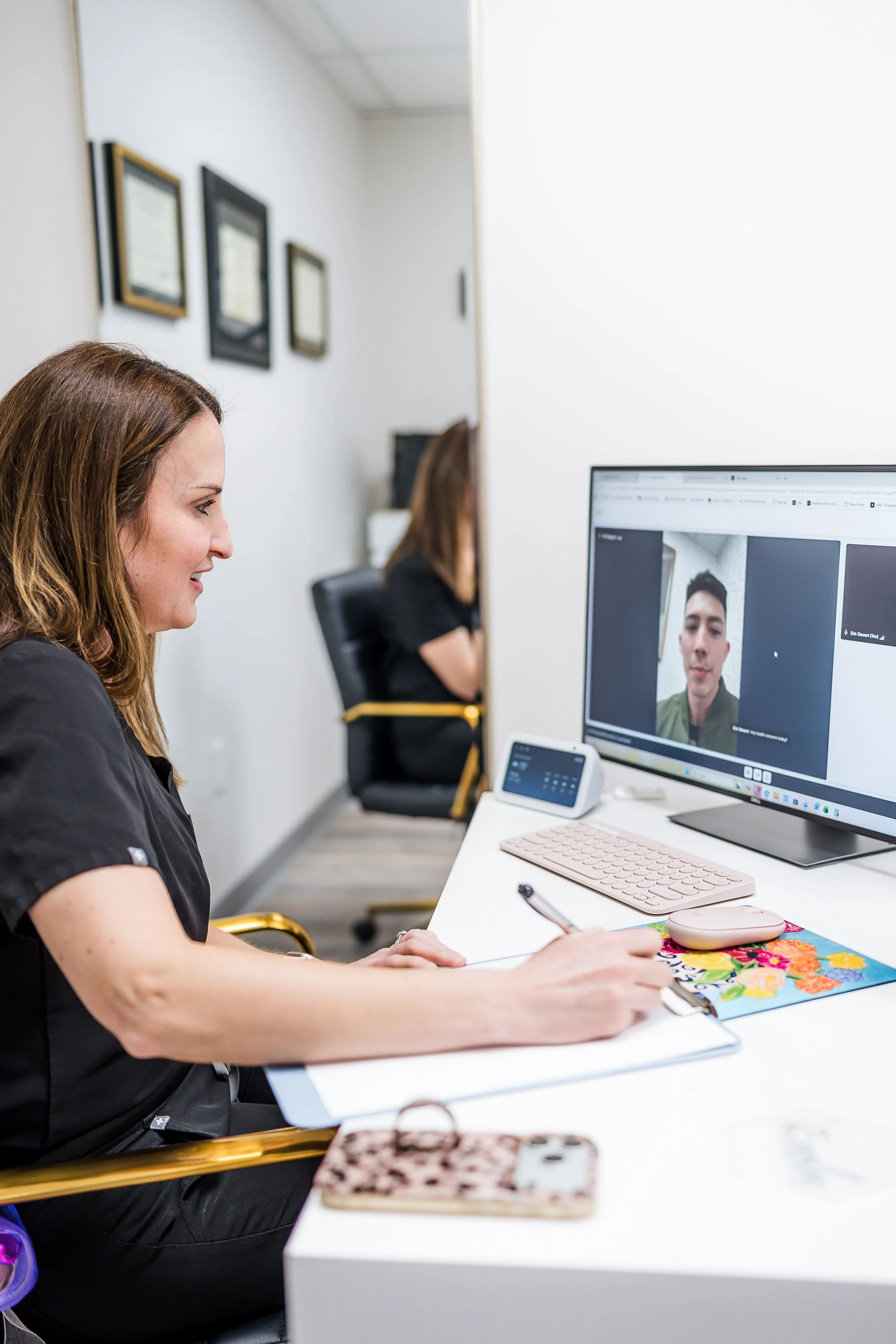 A woman with brown hair smiling while participating in a video call on her computer in an office. She is taking notes on a notepad, with a colorful floral notebook and a pink wireless keyboard and mouse on the desk. Another person is visible in the background, also working at a desk.