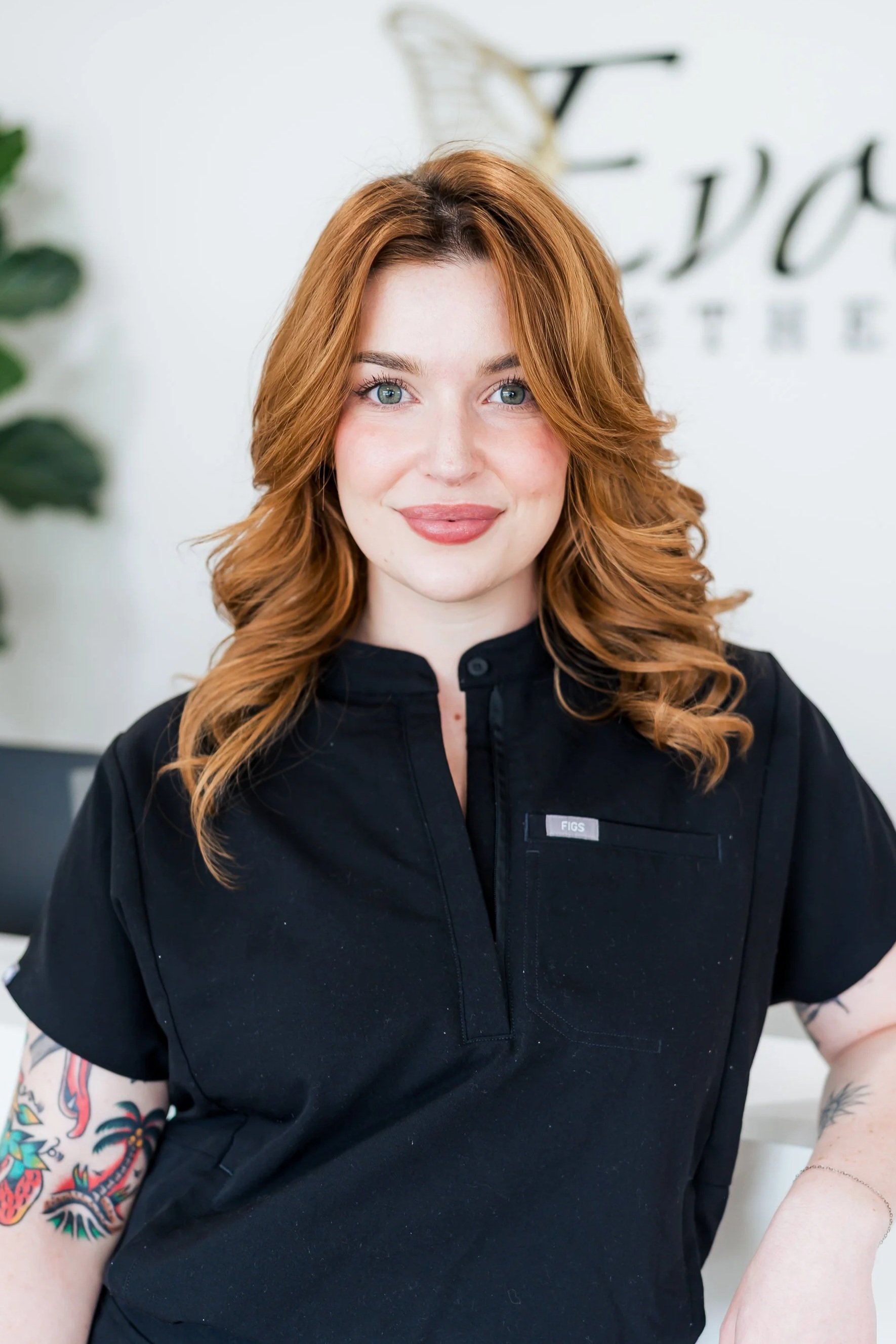 Close-up of a woman with red hair, blue eyes, and tattoos on her arms, wearing a black uniform with a name tag that says 'FIGS', standing in a bright, modern workspace.