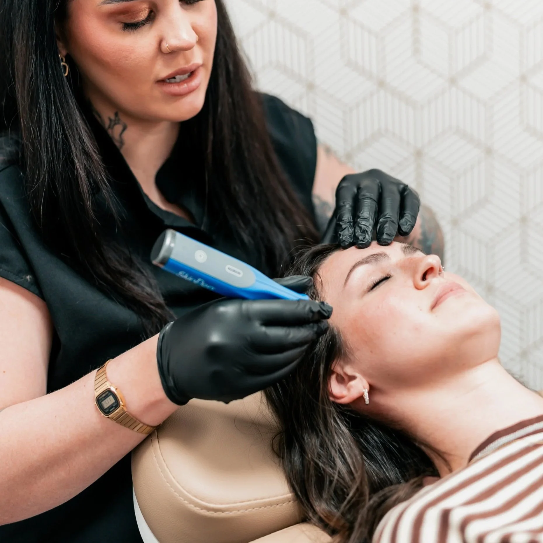 A woman receiving a cosmetic procedure on her face from a technician using a handheld device. The technician is wearing black gloves and appears to be focused on the procedure.