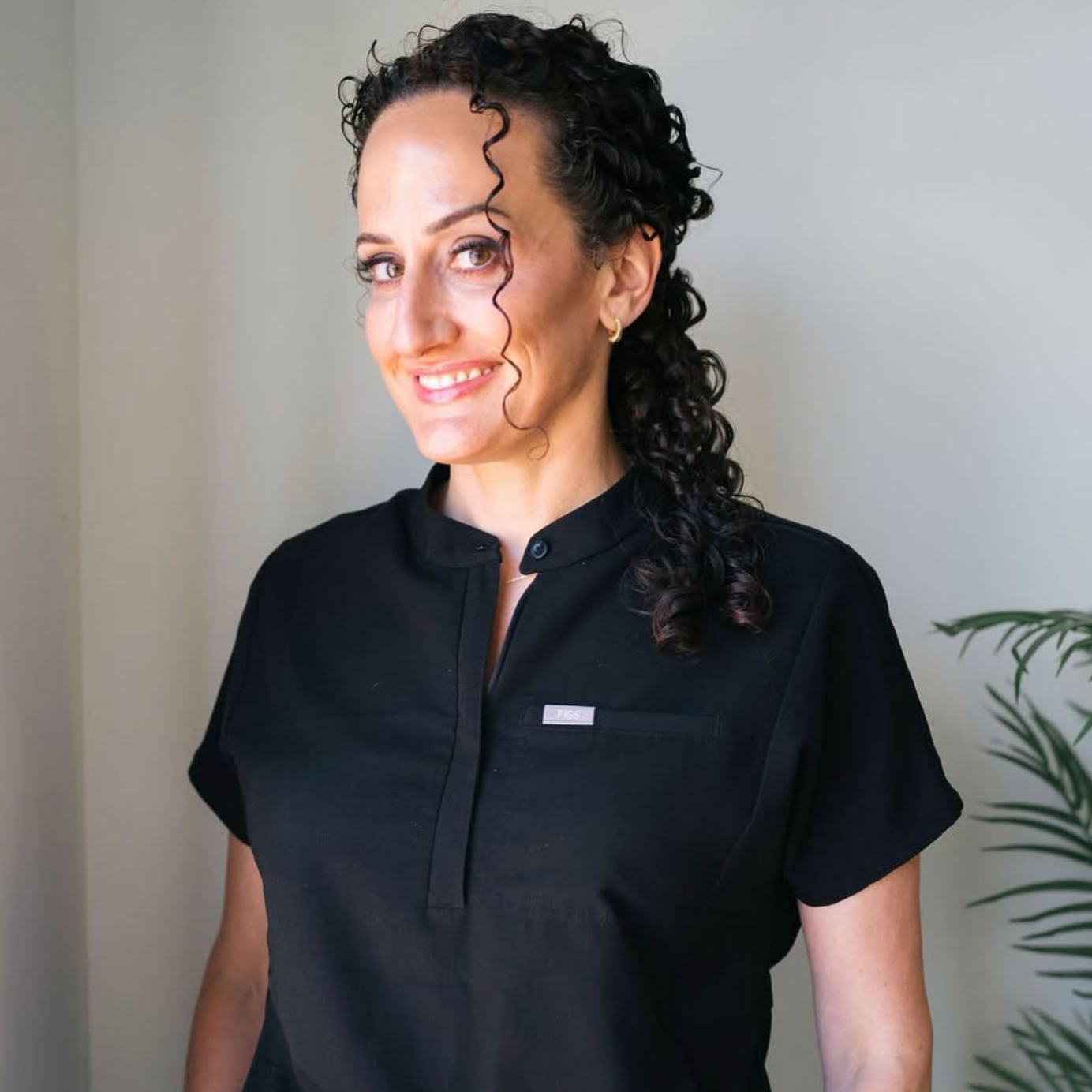 A woman with dark curly hair, wearing a black short-sleeved shirt with a small logo, smiling and looking at the camera, standing indoors near a light-colored wall and a plant.