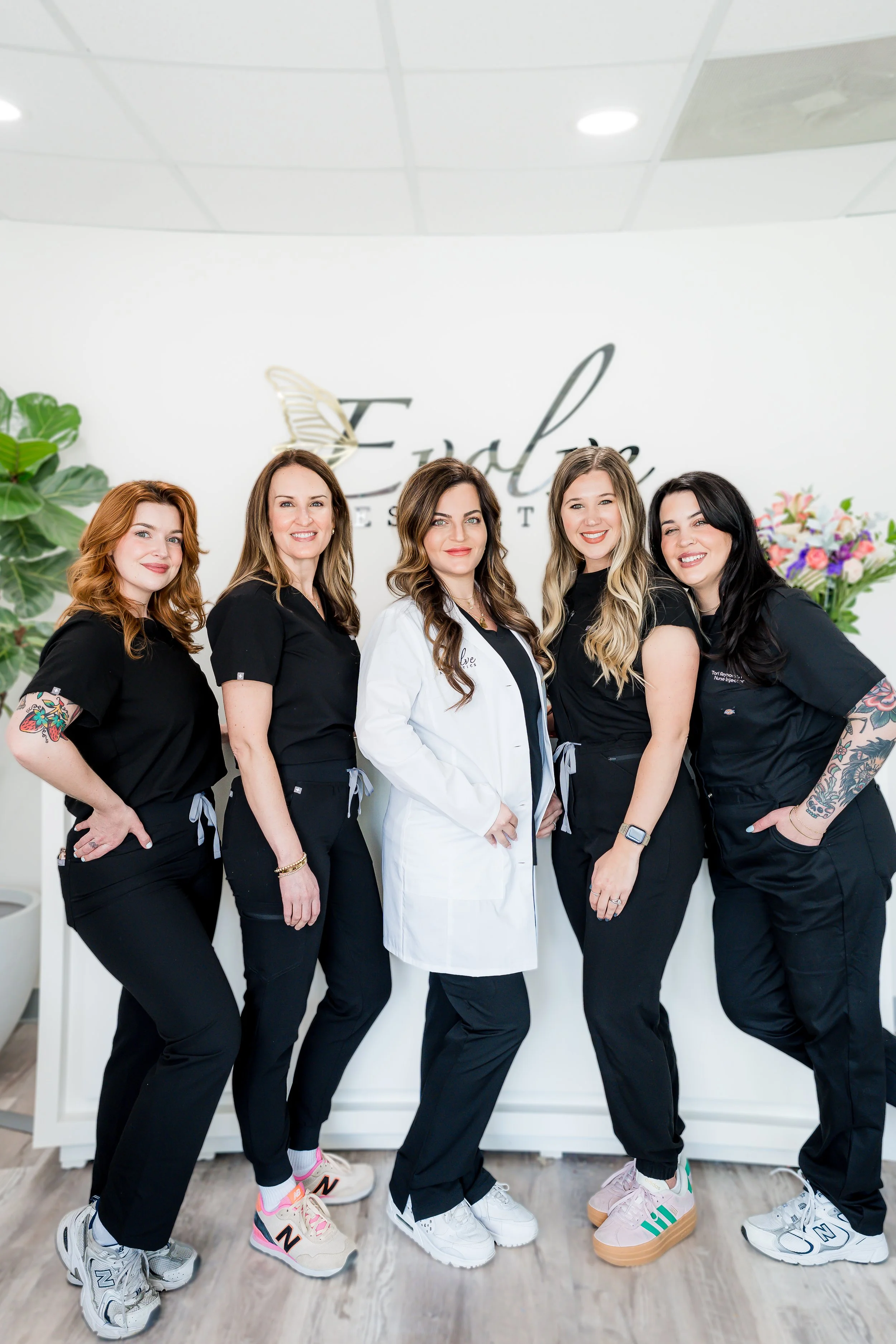 A group of five women in medical scrubs, standing together in a clinic or medical office, with a large white wall behind them and a logo that says "Fable" on the wall, along with some flowers on the side.