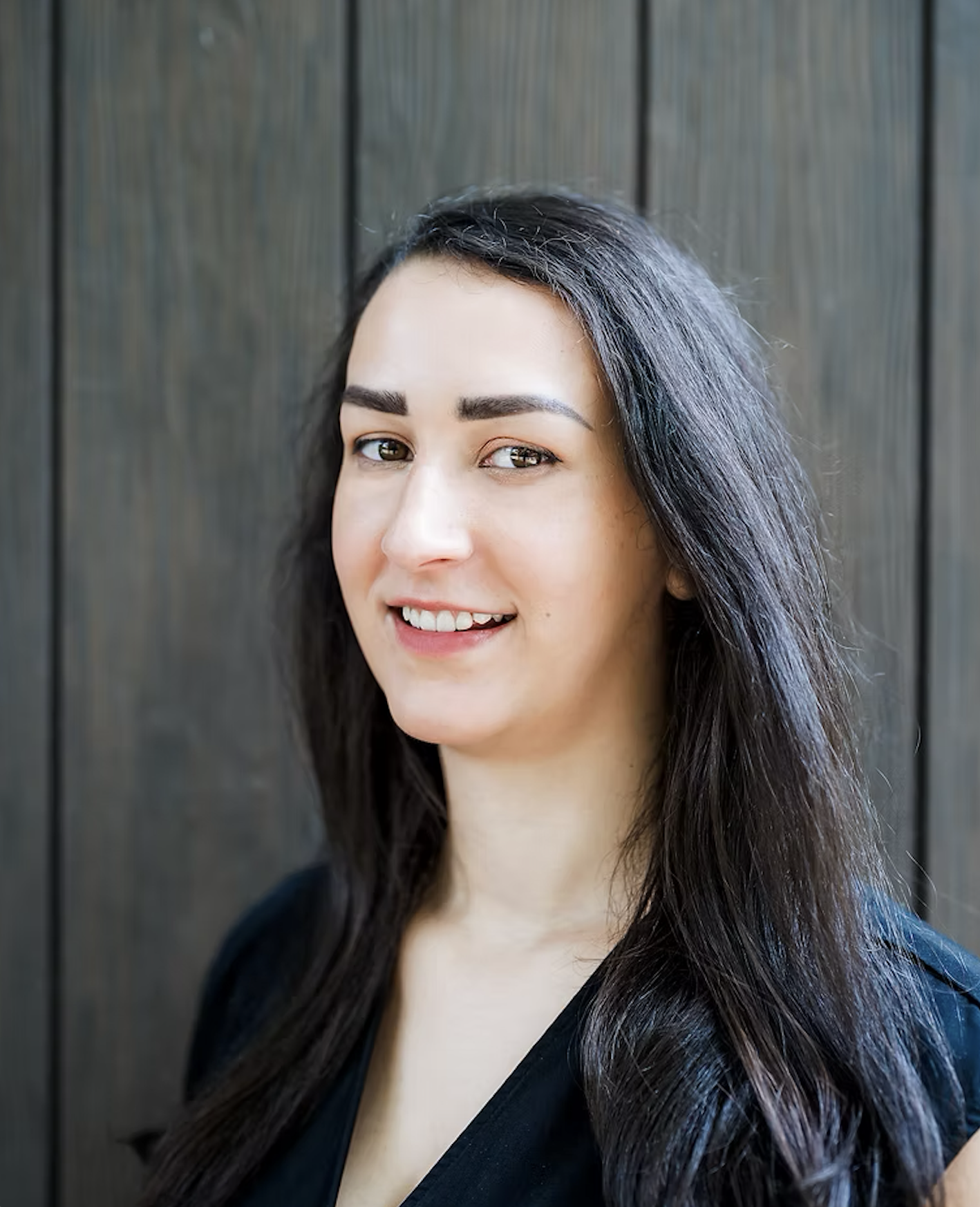 A woman with dark, wavy hair and fair skin smiles softly at the camera, standing in front of a wooden background.