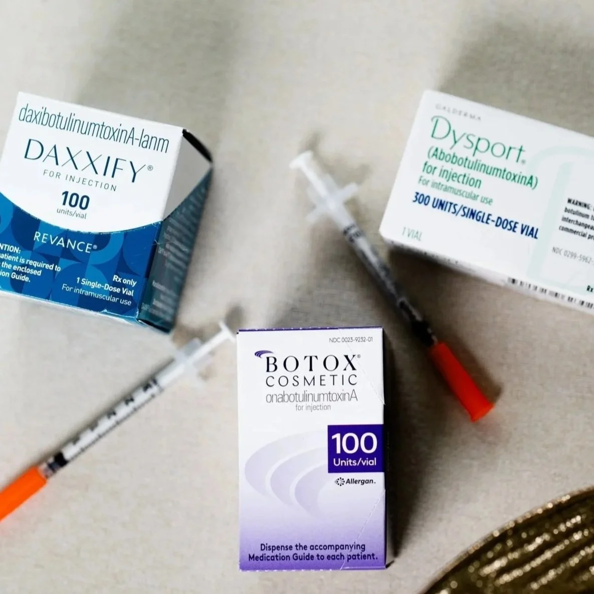 Three boxes of injectable medications, two syringes with orange caps, on a flat surface. The boxes include DAXIFY, Dysport, and Botox.
