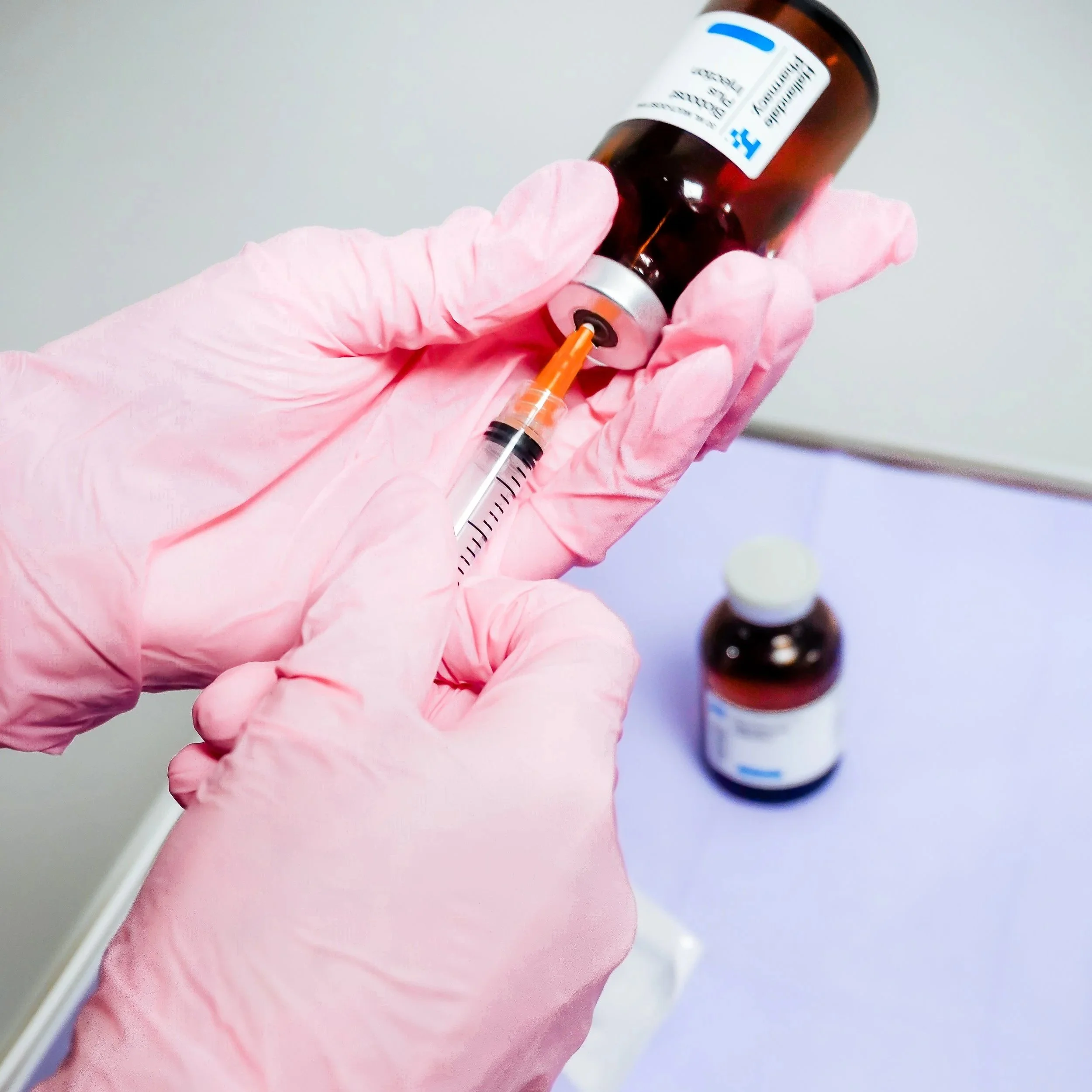 A healthcare worker wearing pink gloves drawing liquid from a brown medicine bottle into a syringe, with another medicine bottle on a light purple surface.