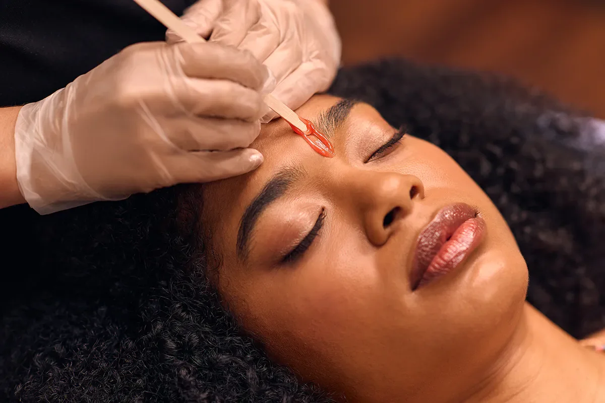 A woman lying down with her eyes closed as a professional applies a red facial treatment to her forehead using a small brush.