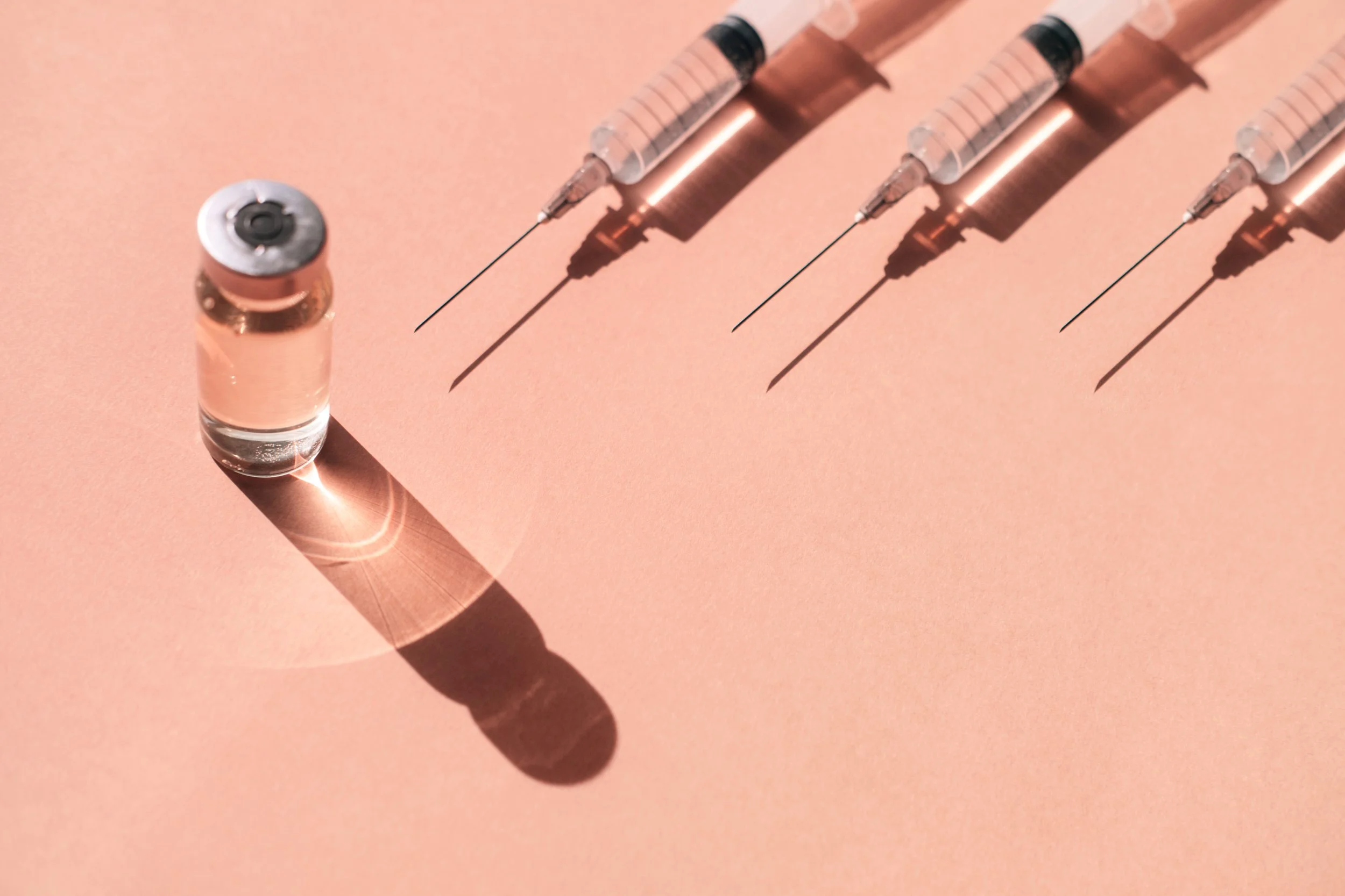 Four botox, daxxify, filler, dysport syringes with needles, one small vial of injection medication, all placed on a peach-colored surface, casting shadows.
