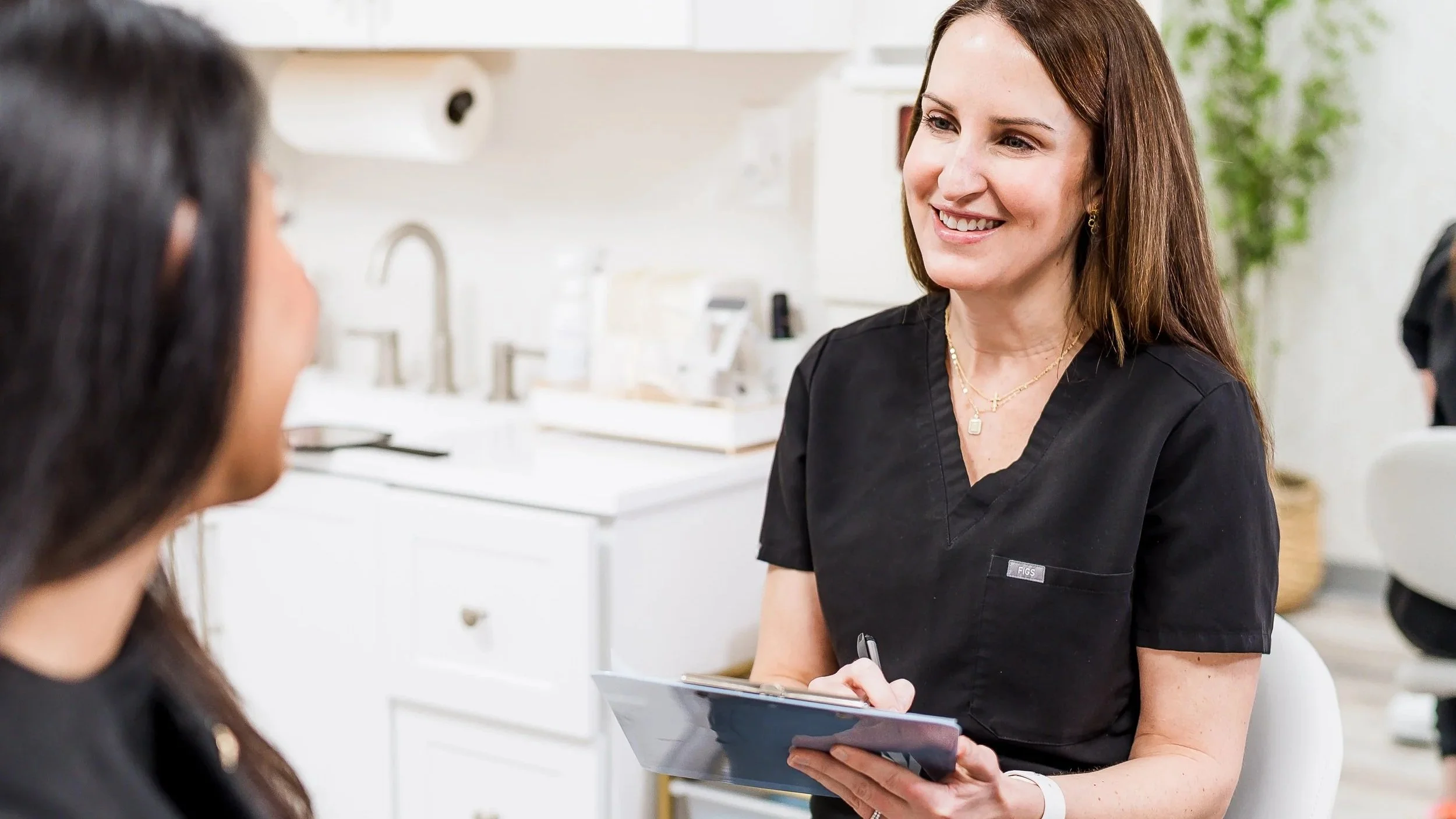 Medical professional in black scrubs smiling and holding a clipboard, interviewing or consulting with a woman with long dark hair in a bright, modern medical office.