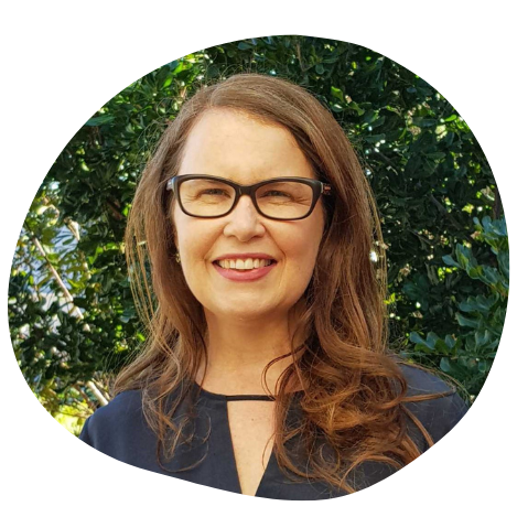 A woman with brown hair, glasses, and a big smile, standing outdoors with green foliage in the background.