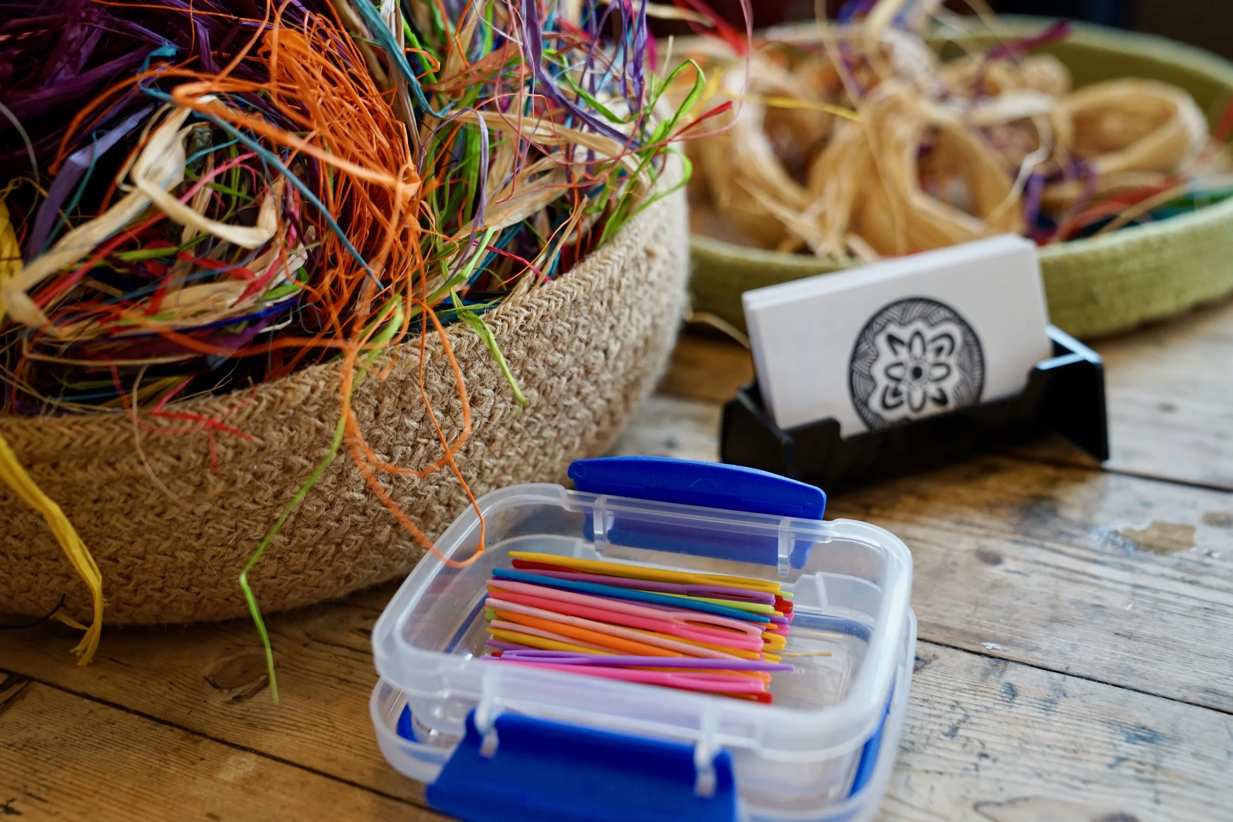 Colorful plastic craft sticks inside a transparent container with a blue lid, set on a wooden surface. In the background, there are baskets filled with shredded paper or raffia, one of which has a cardholder with a card showing a floral design.