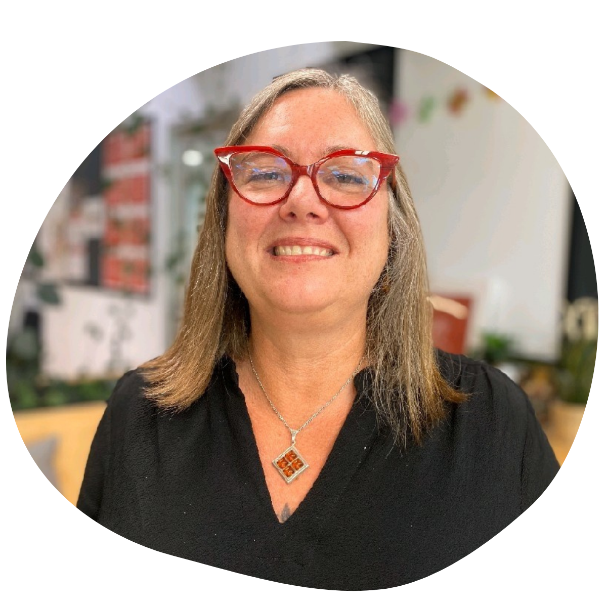 Portrait of a smiling woman with shoulder-length grey hair wearing red cat-eye glasses, a black blouse and a silver necklace with an amber pendant, standing in a bright office with a softly blurred background.
