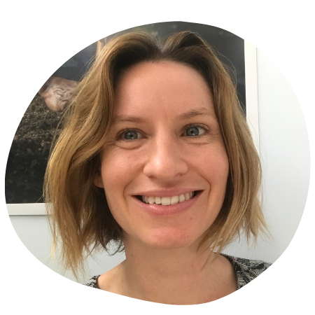 A smiling woman with shoulder-length blonde hair in an indoor setting with artwork behind her.