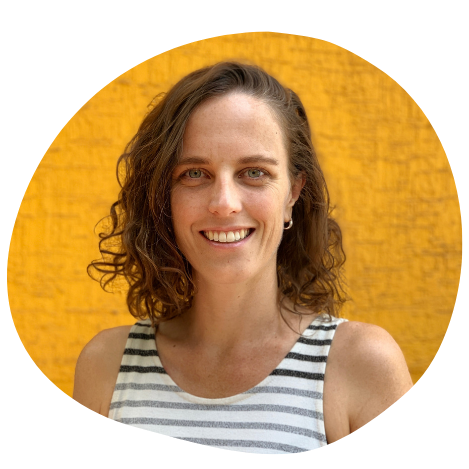 A smiling woman with curly brown hair in front of a yellow wall, wearing a striped sleeveless top.