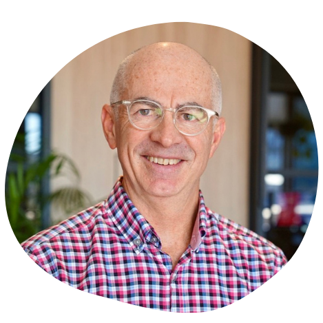 A smiling older man with glasses and a bald head, wearing a checkered shirt, standing indoors near a potted plant.