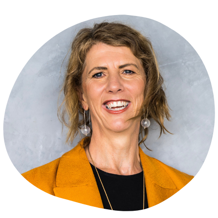 Headshot of a smiling woman with wavy, shoulder-length light brown hair, wearing a mustard yellow blazer and silver earrings, against a textured gray background.