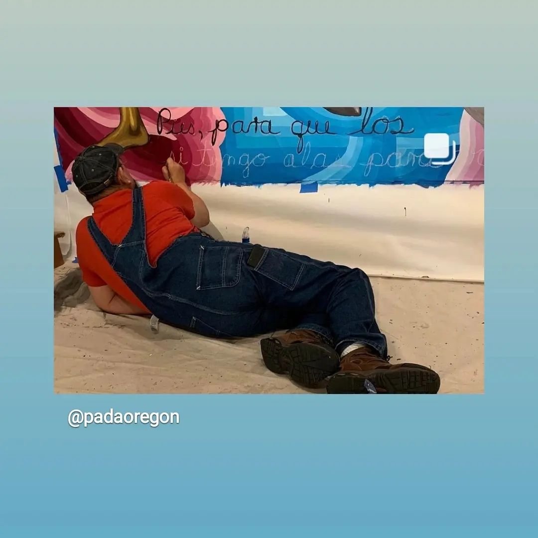 A person lying on the floor, drawing or adding to a large colorful mural on a wall.