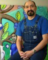 A man with a beard wearing a blue shirt and apron standing in front of a colorful mural with leaves and flowers.