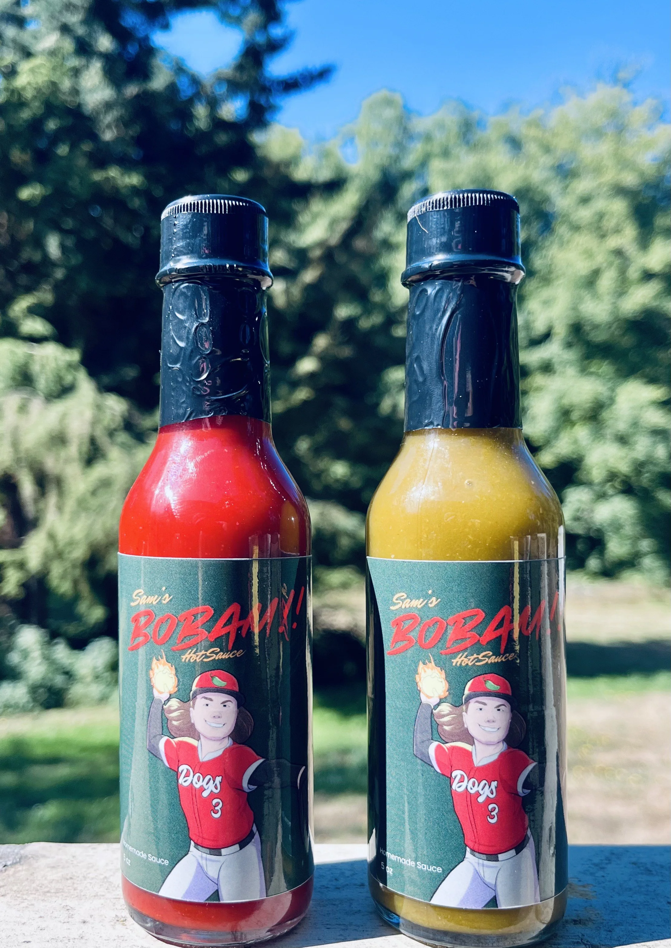 Two bottles of hot sauce labeled 'Sam's BOMBA! Hot Sauce,' one red and one yellow. Each bottle has a label featuring a cartoon woman in a baseball uniform, with a flame in her hand. The background is outdoors with trees and a blue sky.