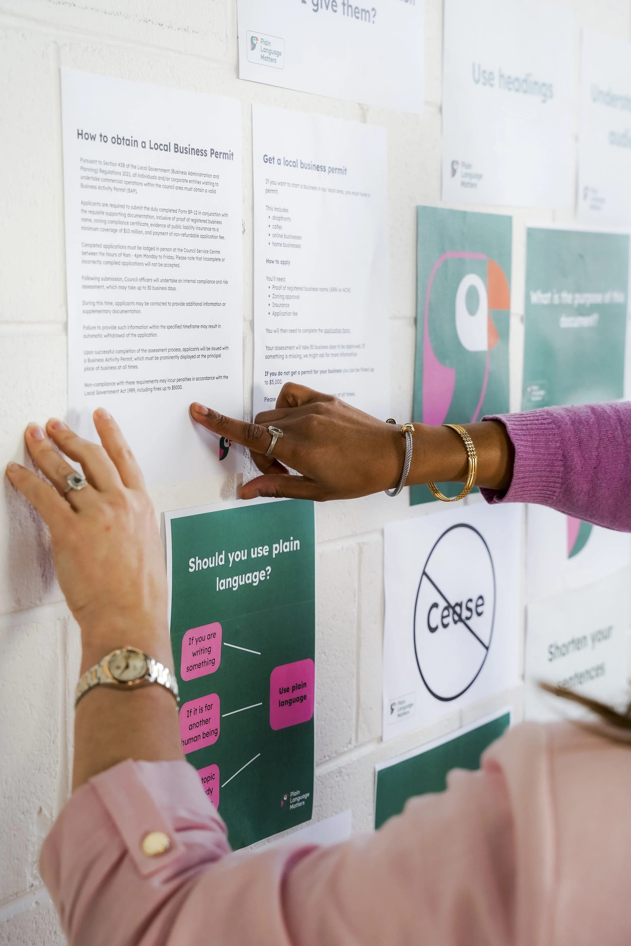 Two people putting up a poster about whether to use plain language or not, with other informational papers on a wall.