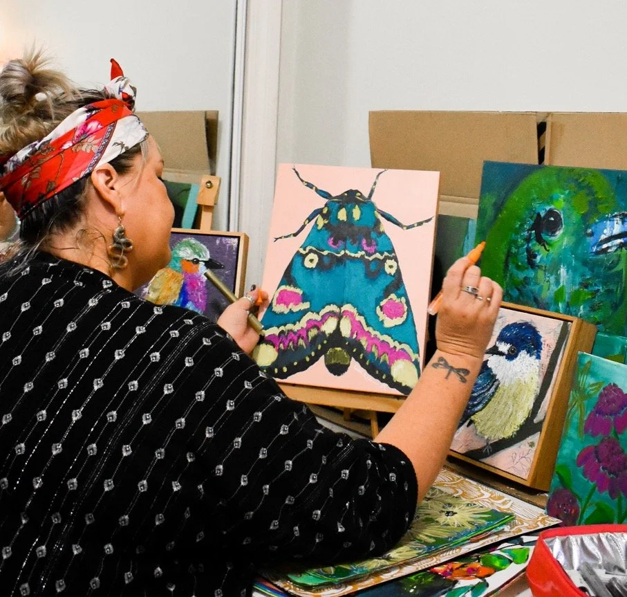 A woman with a red bandana and earrings painting a colorful moth on a canvas. The area is filled with several paintings, including birds and abstract designs, at an art display or studio.