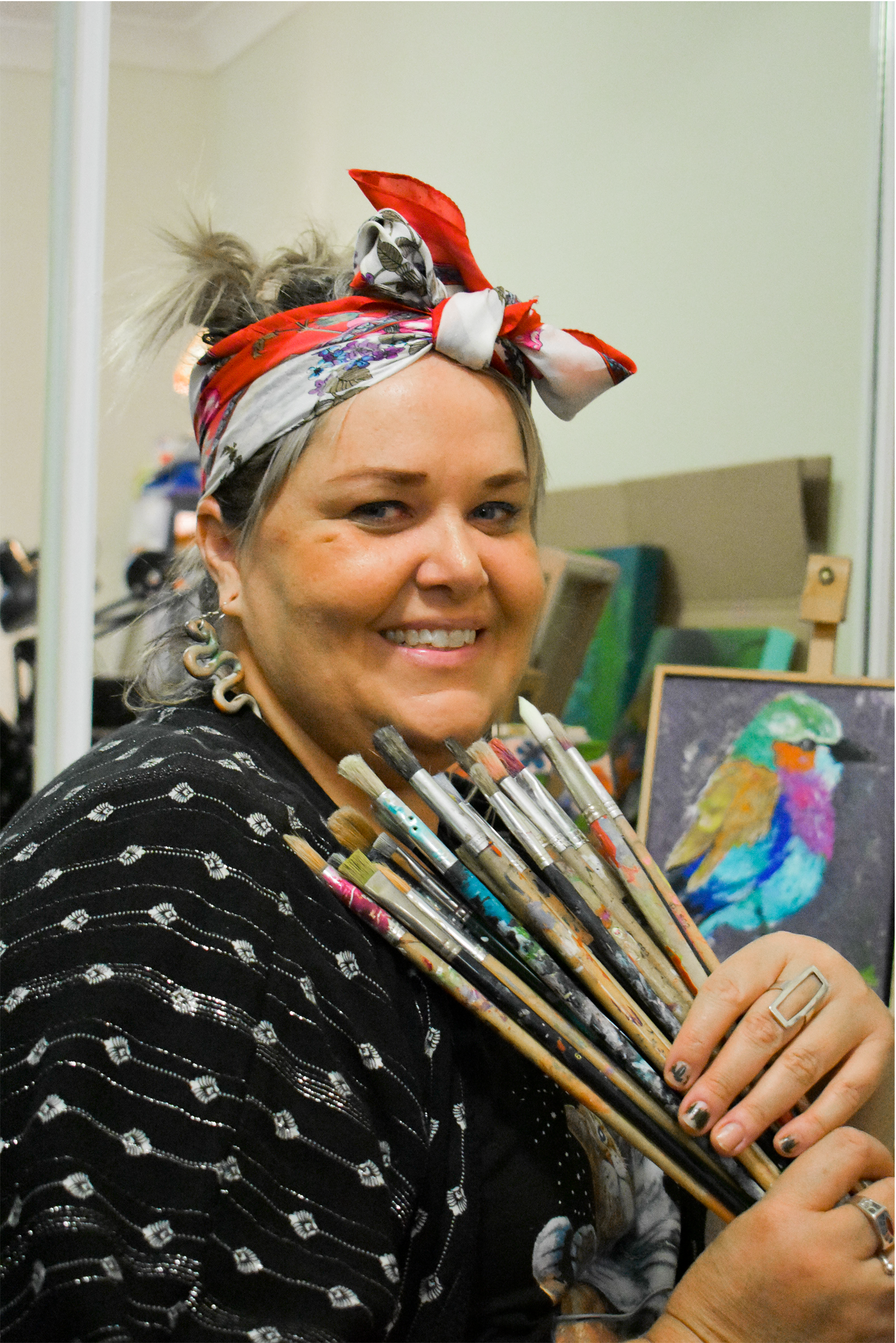 A woman smiling and holding a bunch of paintbrushes, wearing a black patterned top and a red and white headscarf. There is a painting of a colorful bird in the background.