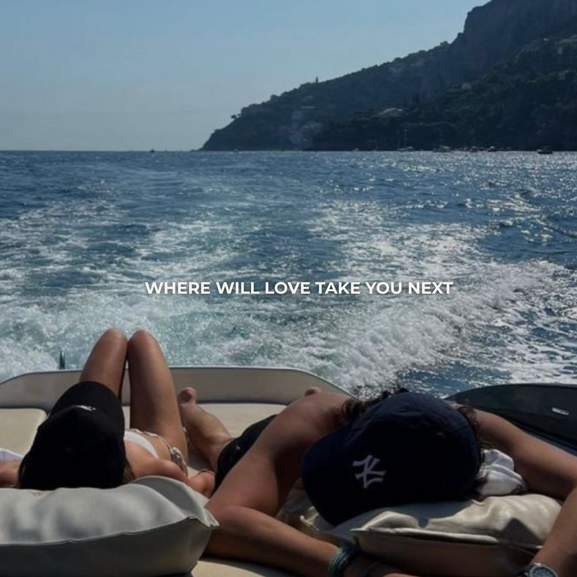 Two people, a man and a woman, lying on a boat seat facing away from the camera with the ocean and a hillside in the background. The man is wearing a navy cap and the woman is in a swimsuit.